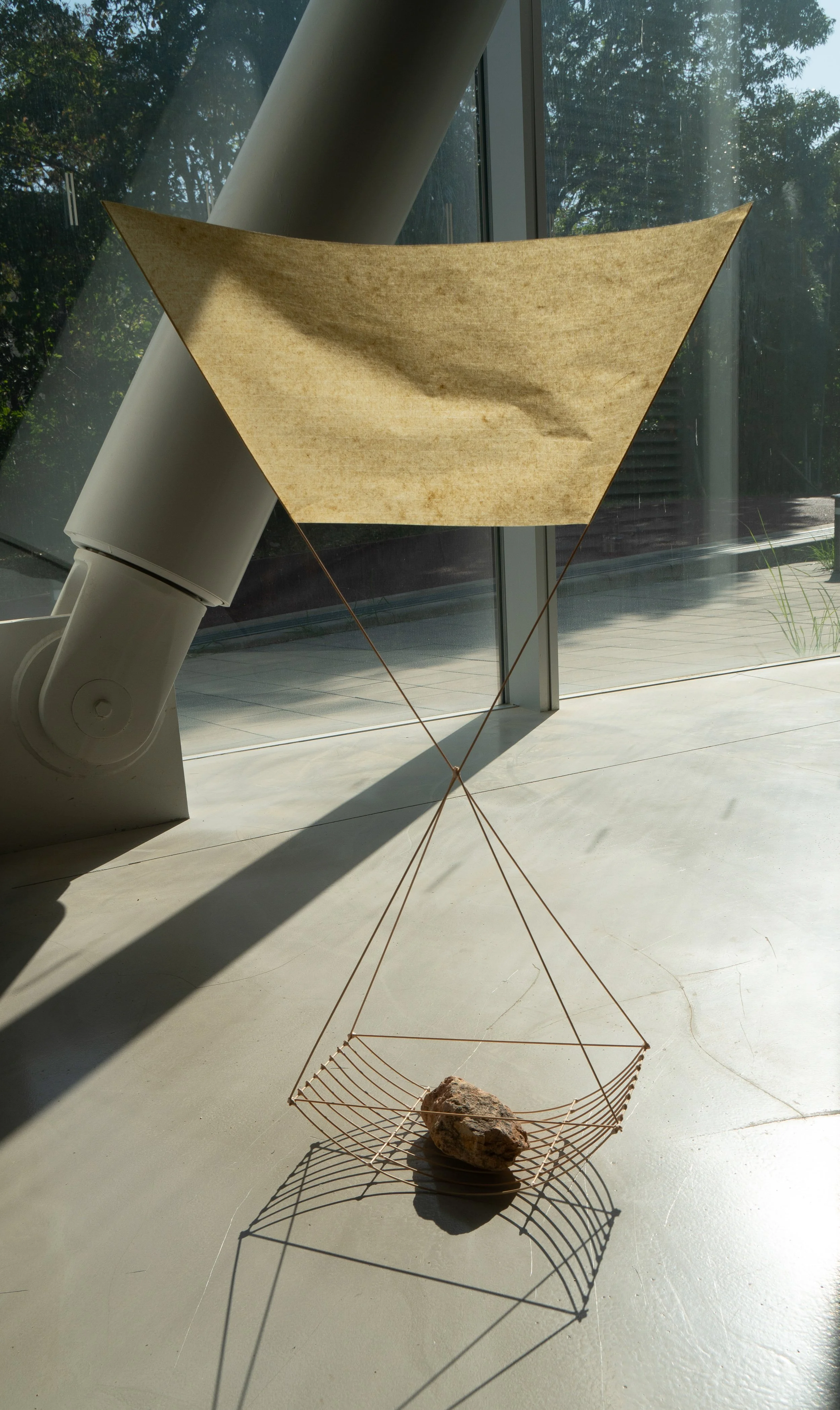 A minimalist art installation featuring a geometric wire platform with a large rock, suspended by strings attached to a beige triangle fabric, all situated near a large window with natural sunlight.