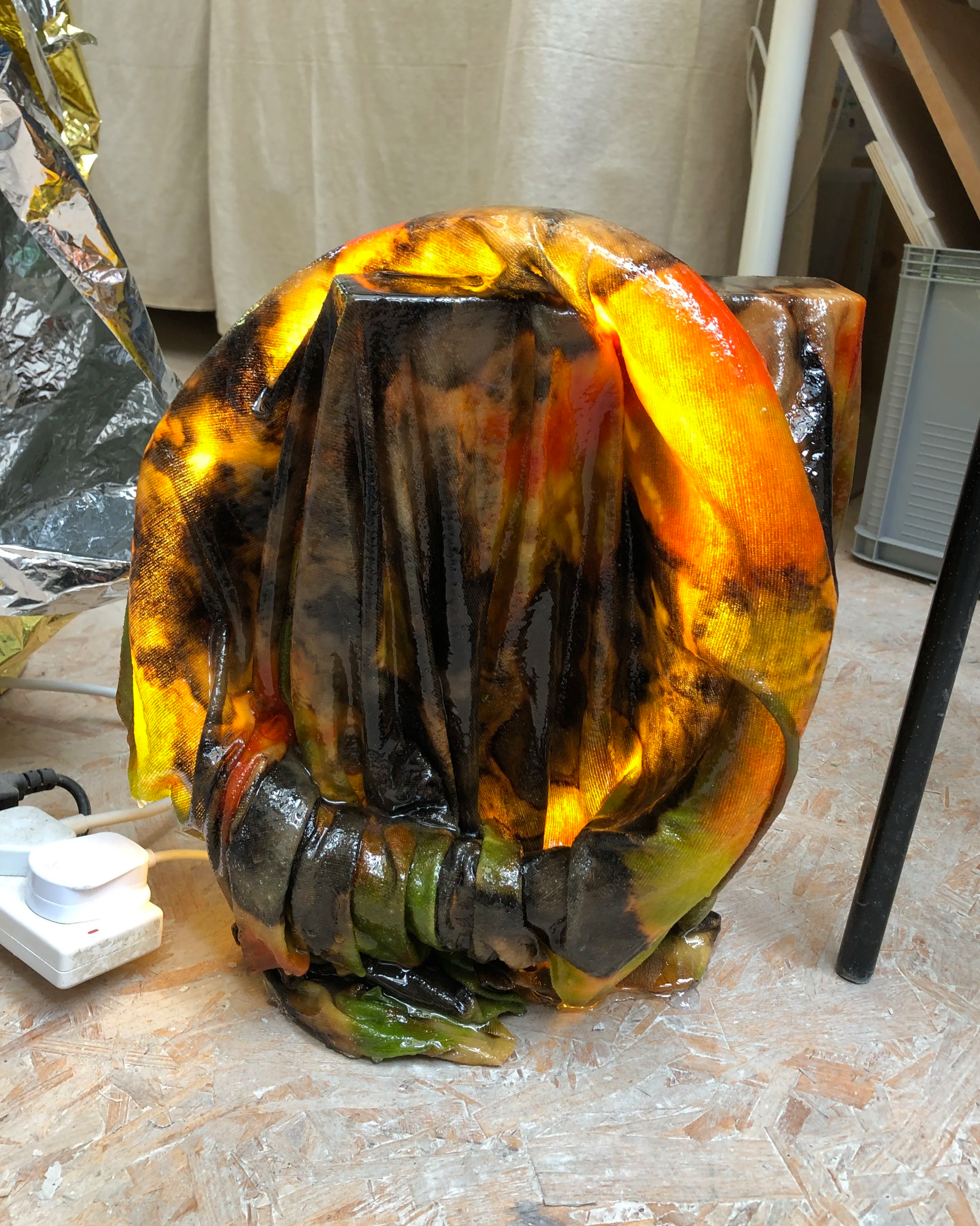 Burned and melted plastic or rubber object, glowing from heat, placed on a wooden surface near an electrical outlet and a metal table leg.