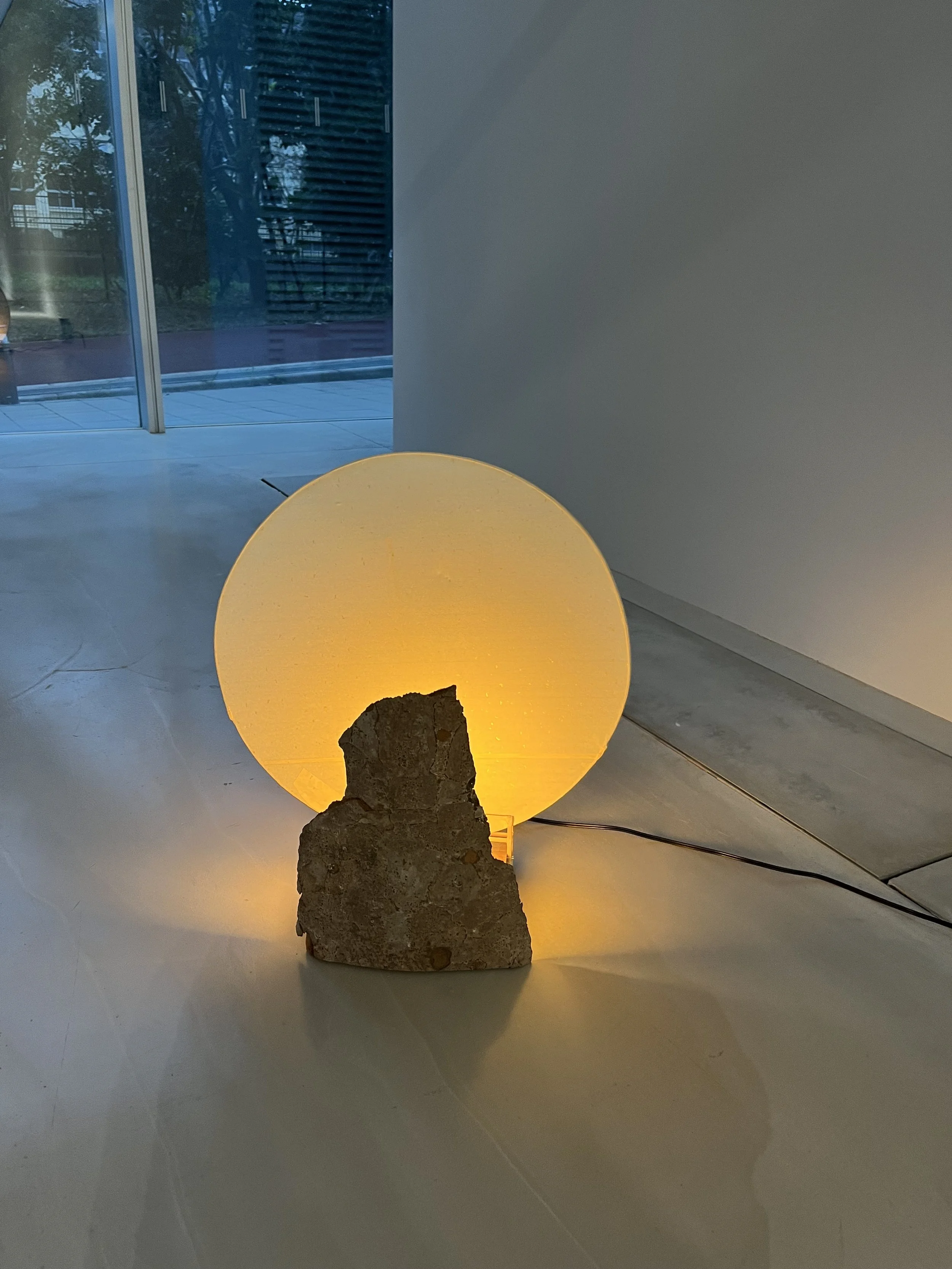 A glowing yellow spherical lamp placed on a flat surface, supported by a rough rock, with an underground electrical cord running from the lamp. The background includes a window reflecting outdoor trees and structures, along with a plain white wall.