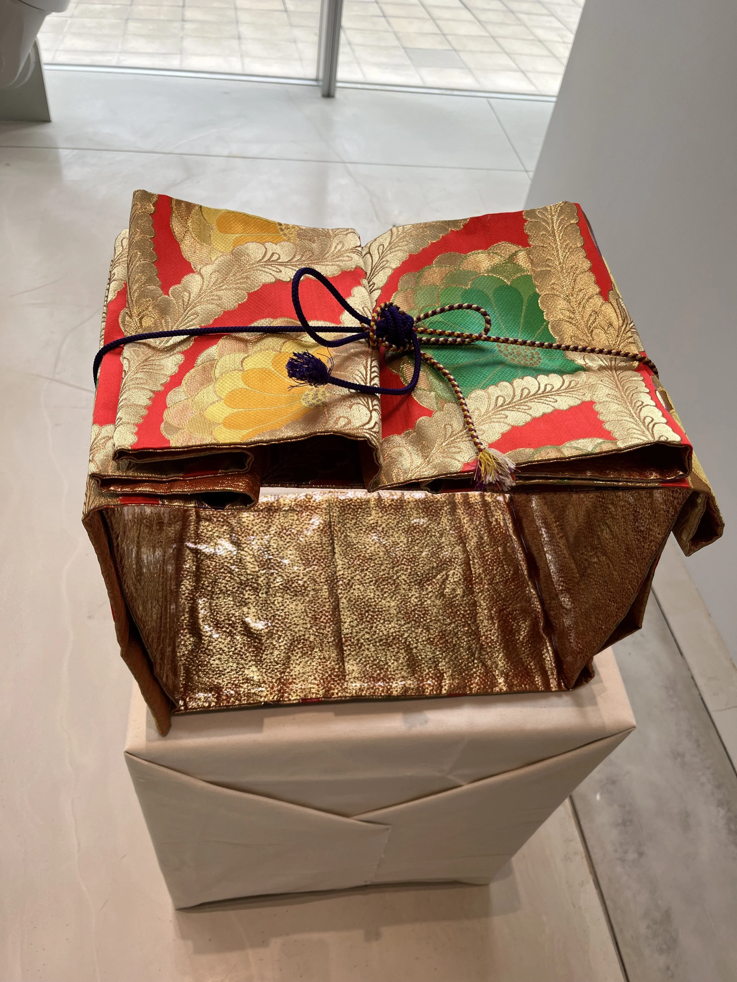 A wrapped gift box with gold and red patterned cloth on top, tied with a decorative purple and gold string, placed on a beige-wrapped box.