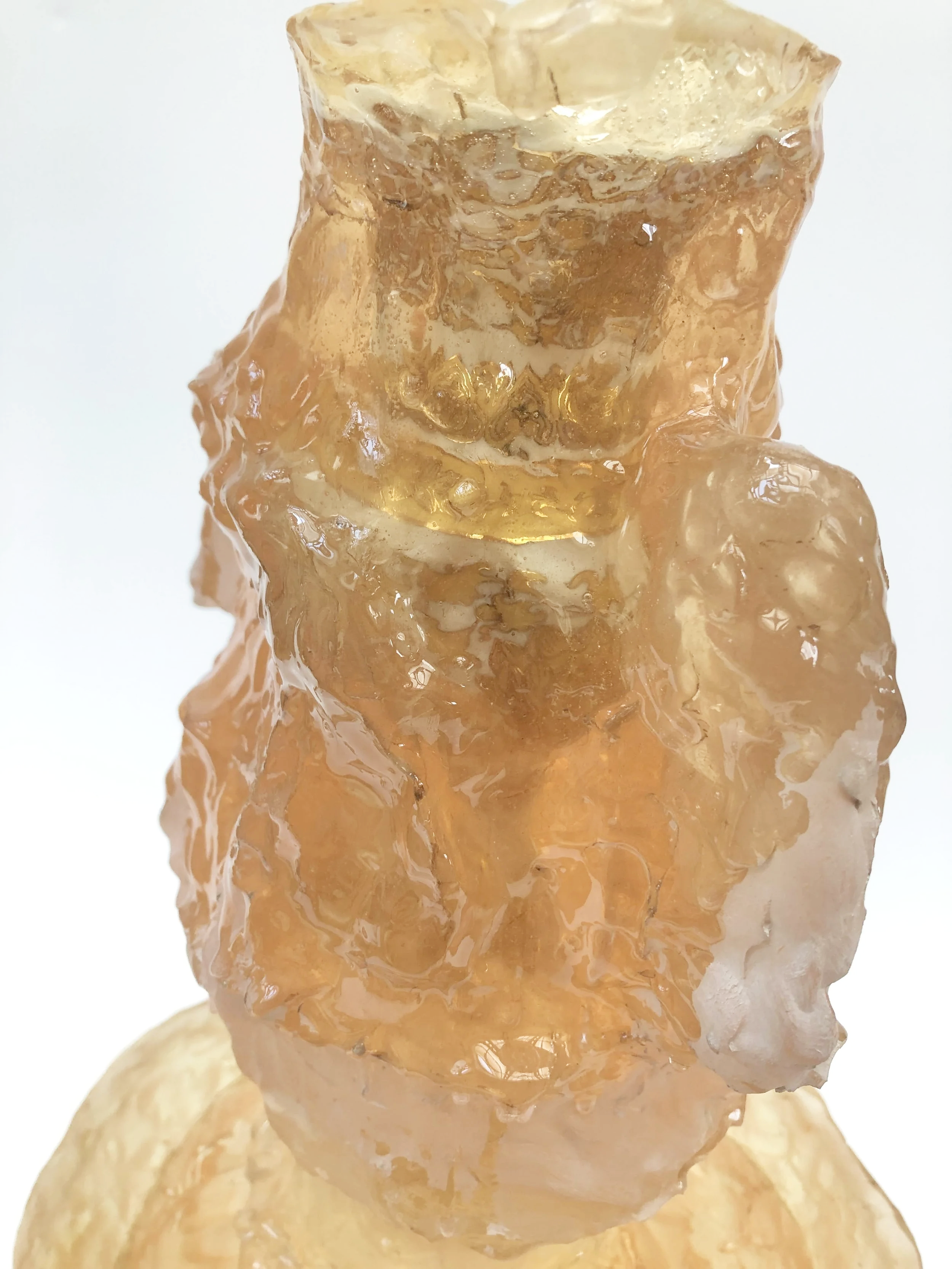 Close-up of an abstract glass sculpture with a rough, textured surface, made of amber-colored and translucent glass, set against a white background.