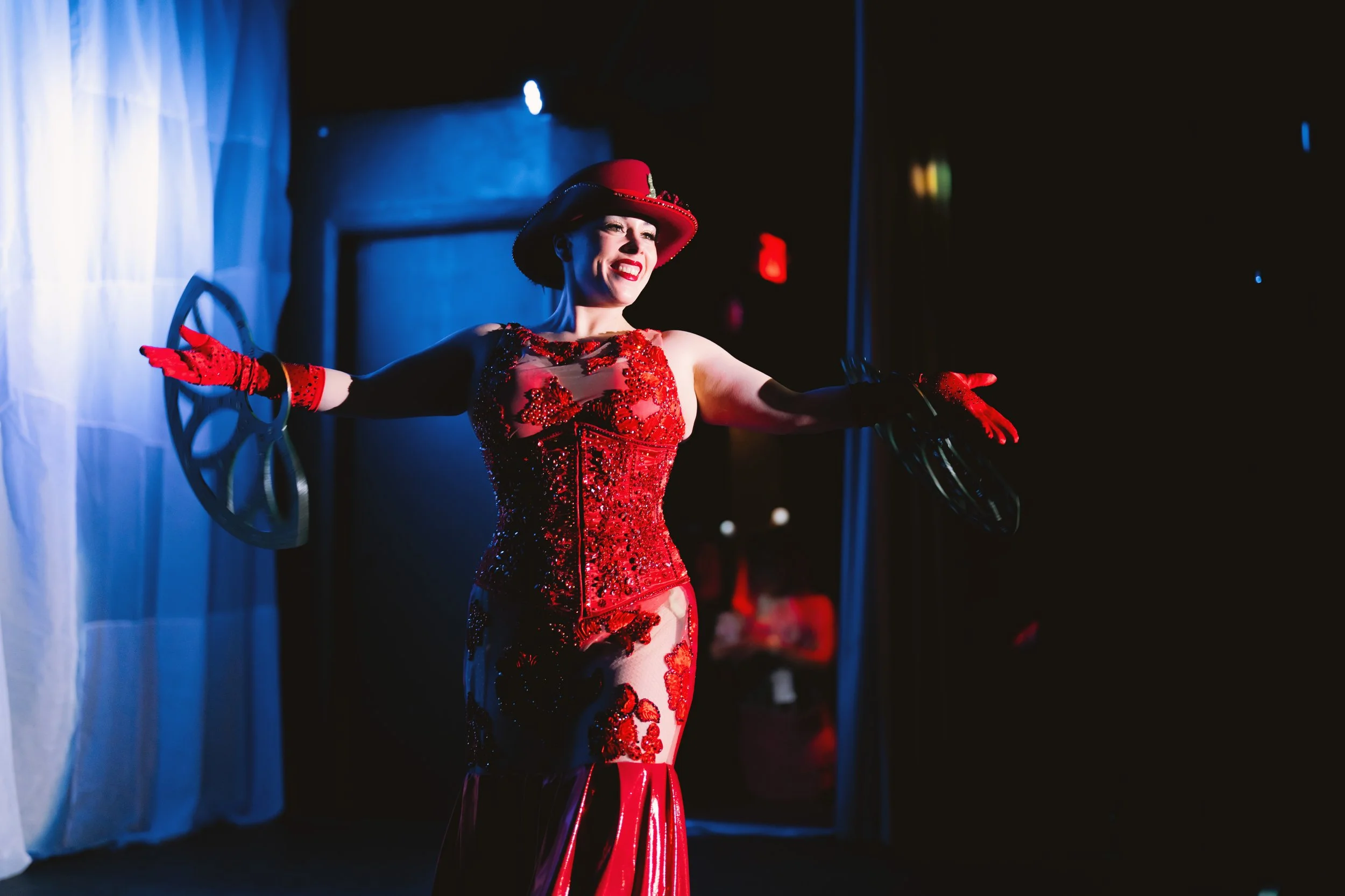A woman in a red hat and a red sequined dress standing on stage with outstretched arms and a big smile.