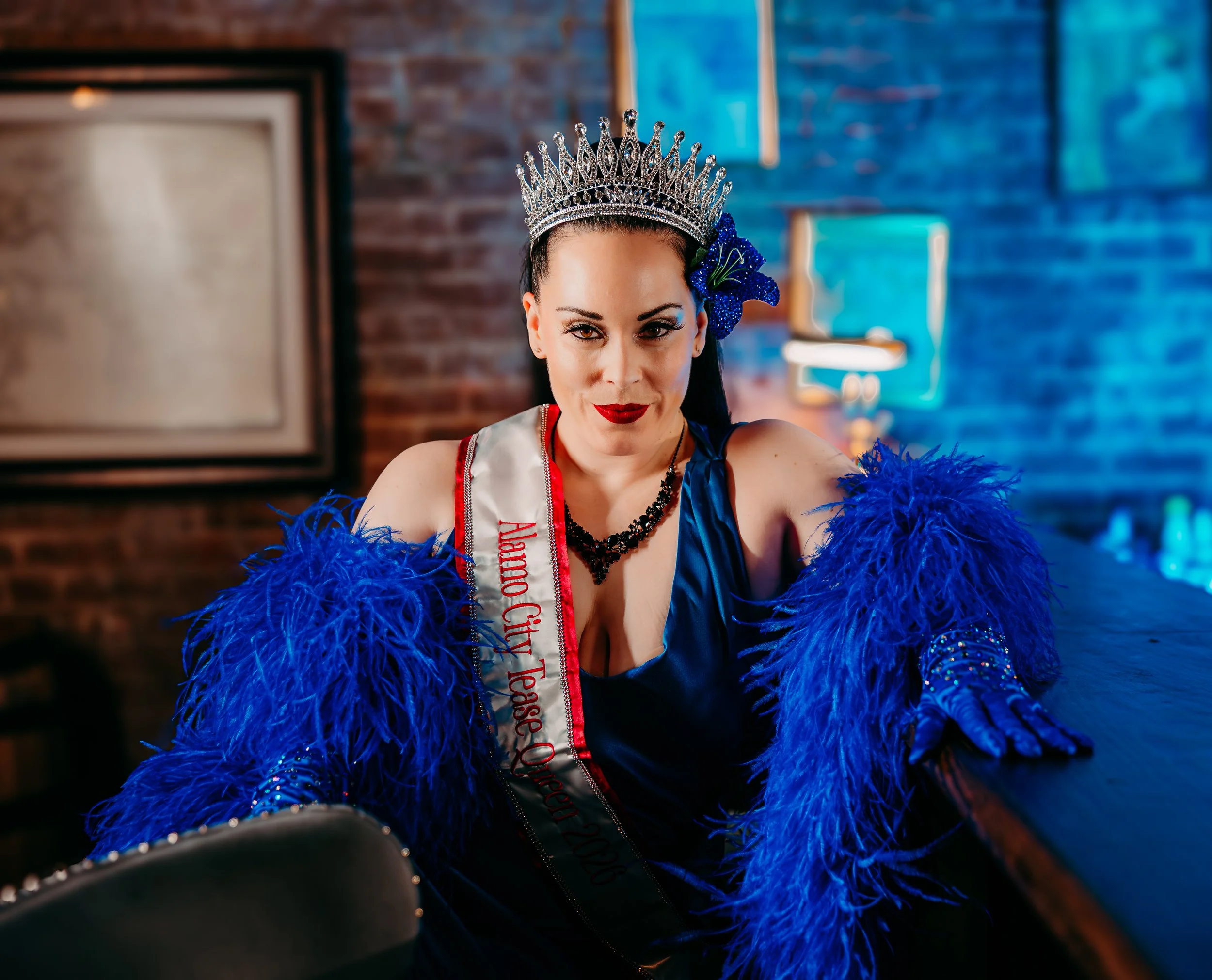 A woman wearing a crown and a sash that reads 'Queen of the City' while sitting at a table with blue feather boas, with a brick wall and a television in the background.