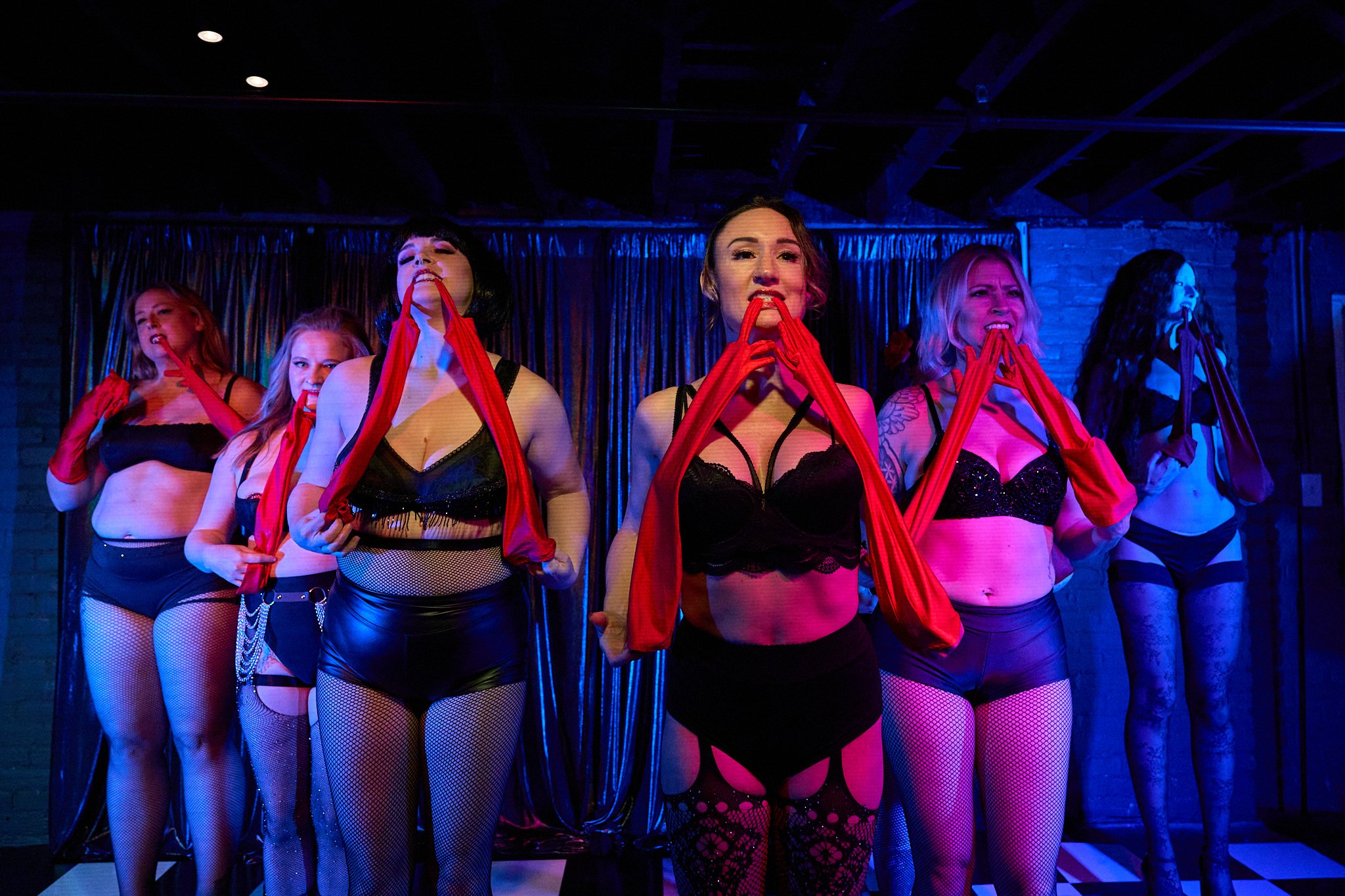Women on stage wearing lingerie, fishnet stockings, and red gloves, performing in a nightclub with a dark background and blue lighting.