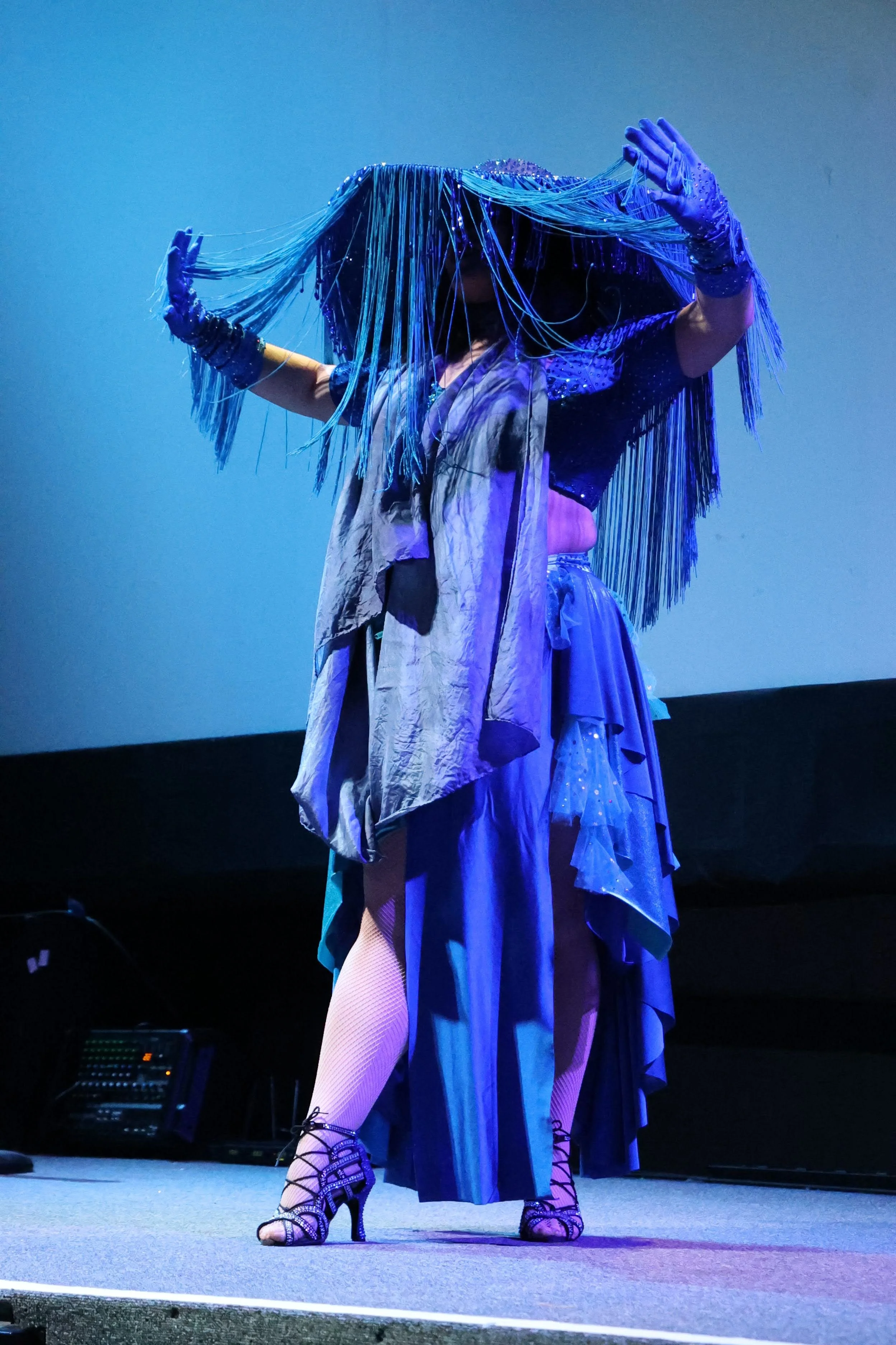 Performer on stage wearing a layered dress, long gloves, and high heels, holding long strands of beads or fringe in both hands, with stage lighting creating a purple and blue hue.