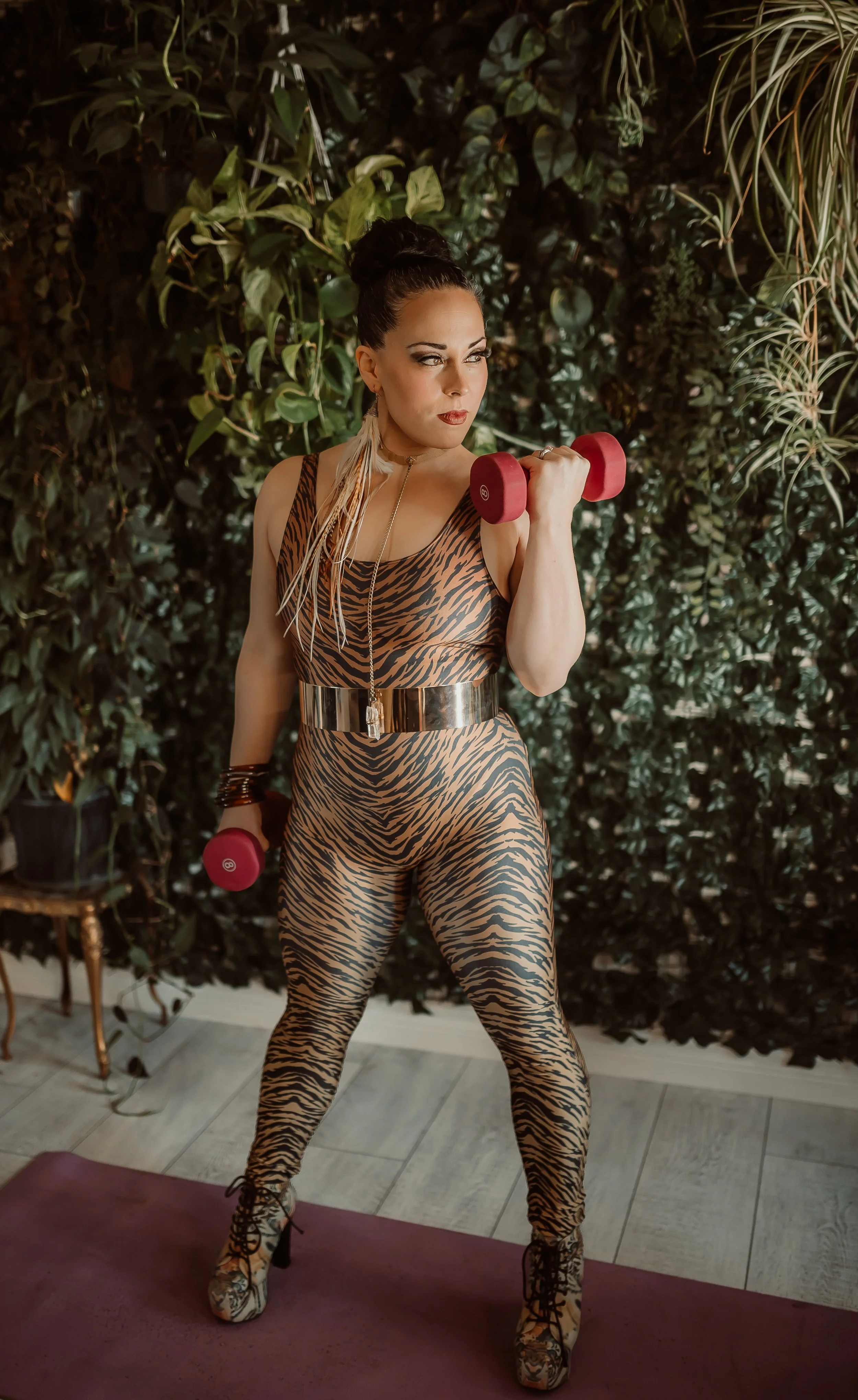 A woman in a tiger print workout outfit lifting pink dumbbells in front of a lush green leafy background.
