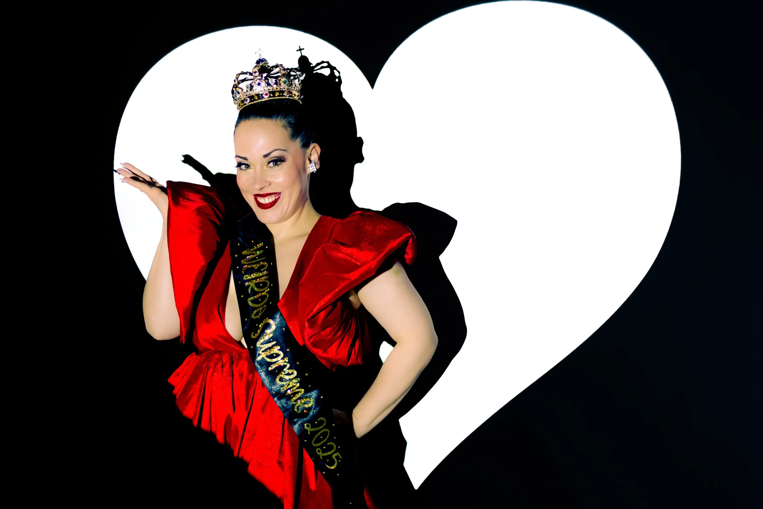 Woman dressed in a red dress with a crown and tiara, wearing a sash. She is smiling and posing with her hand raised, standing in front of a large white heart shape.