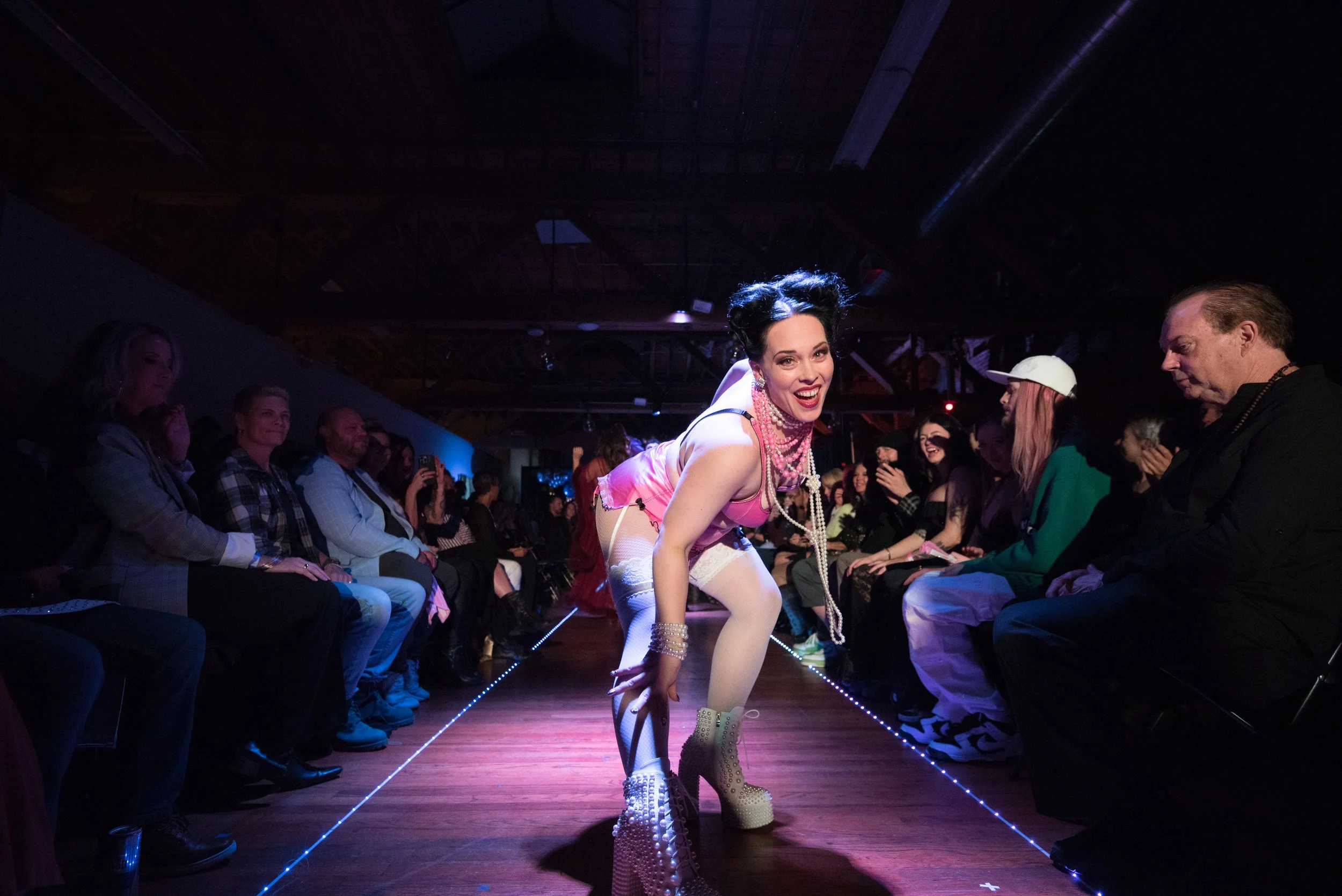 Performer dressed in pink with pearls, high heels, and stockings, performing on a runway with an audience watching in a dimly lit venue.