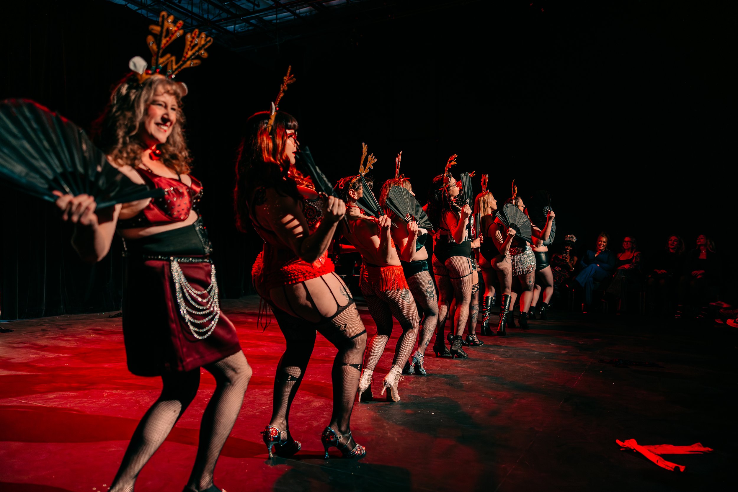 A group of performers dressed in red and black costumes with fishnet stockings and high heels, wearing antler headbands, are lined up on stage holding fans, during a dance performance under stage lighting.