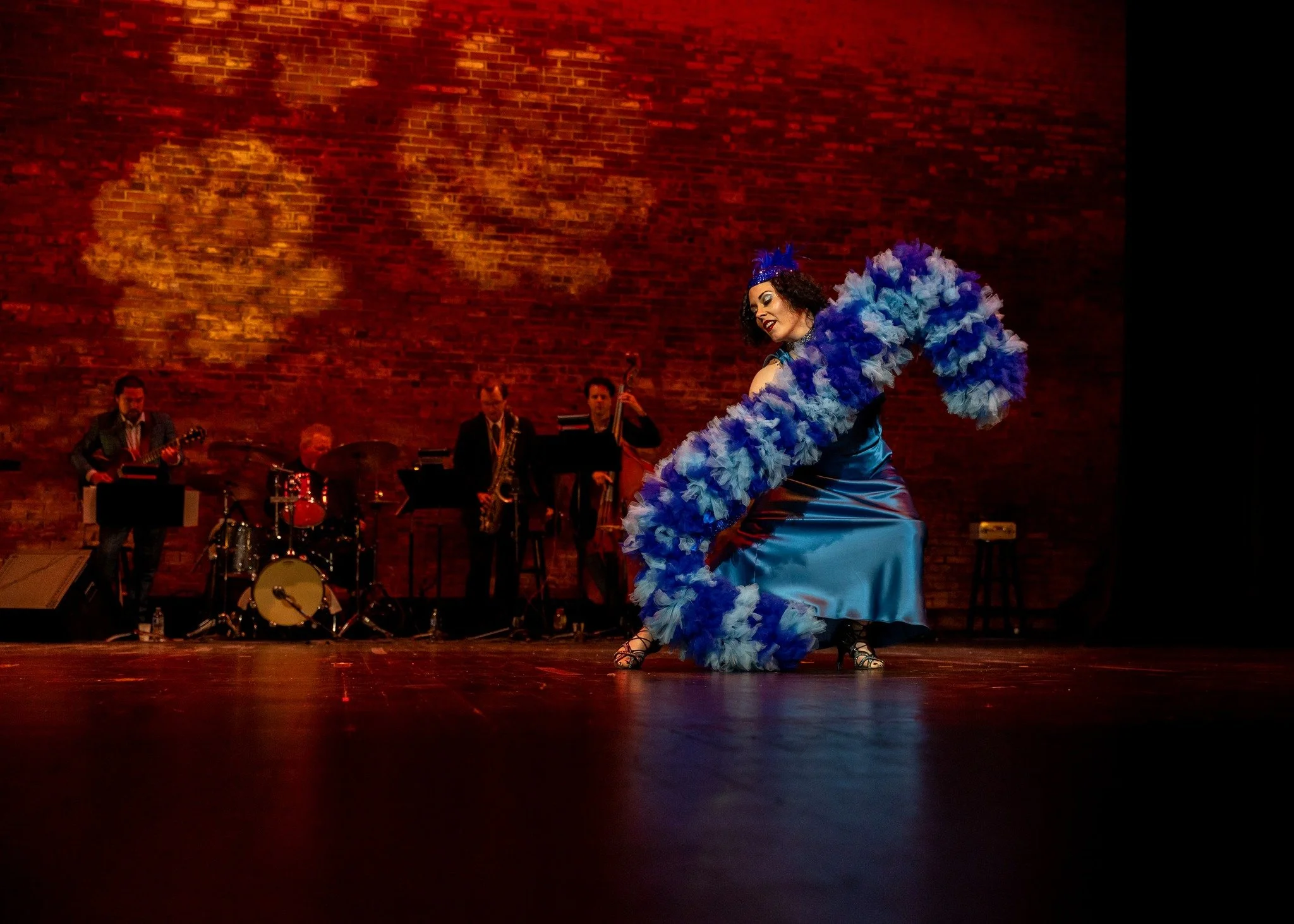 Performer in a blue dress with blue feathered accents dancing on stage with a band playing in the background against a brick wall.