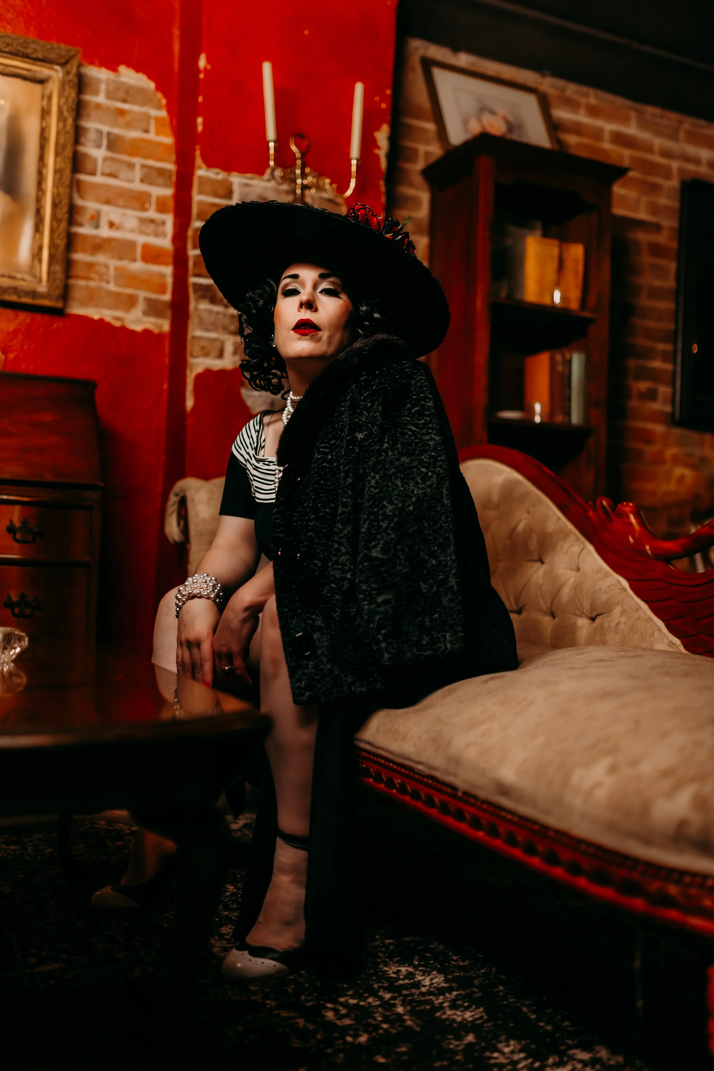 A woman dressed in vintage clothing and jewelry, wearing a large black hat, sits on a patterned sofa in a room with brick walls, a wooden bookshelf, and a gold-framed painting.