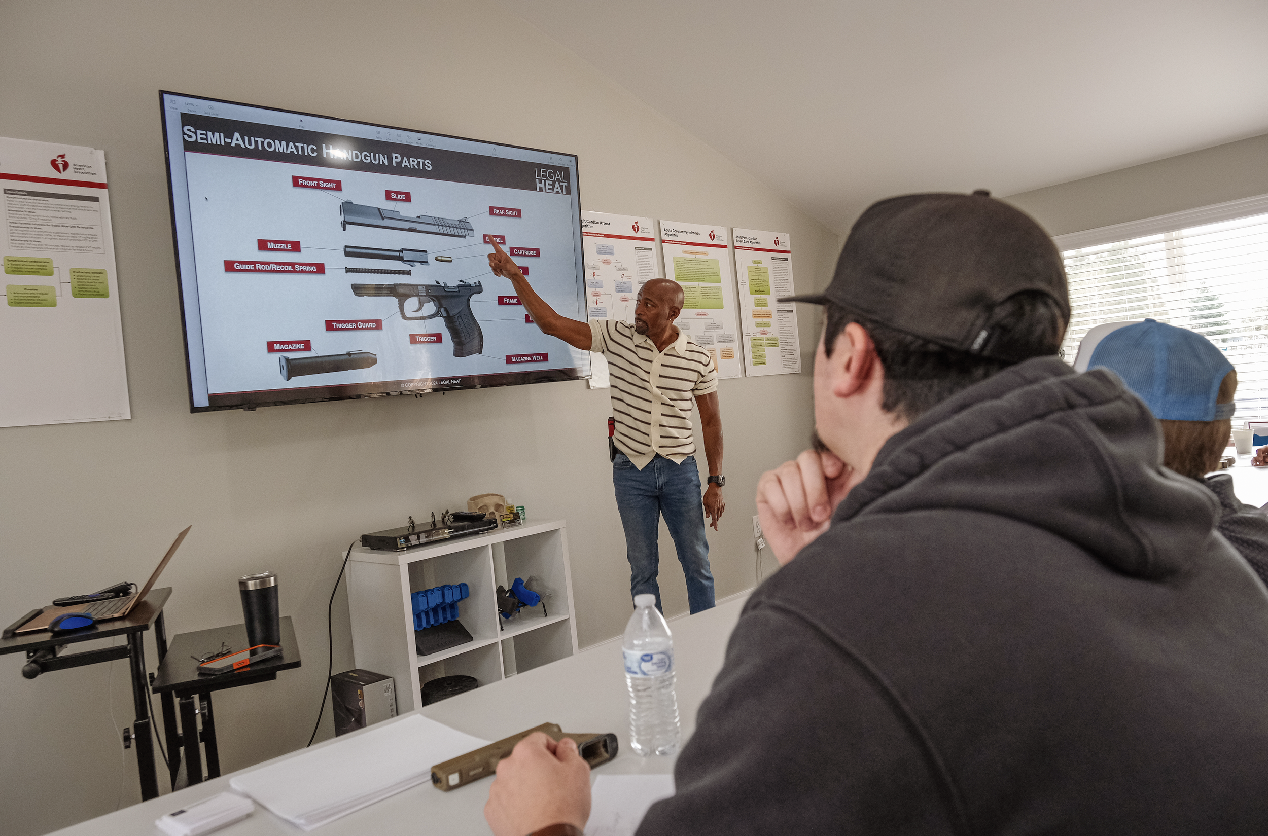 K1 Risk Concealed Handgun License group class in a classroom or training room, with several posters on the wall and a large screen displaying labeled diagram of handgun components.