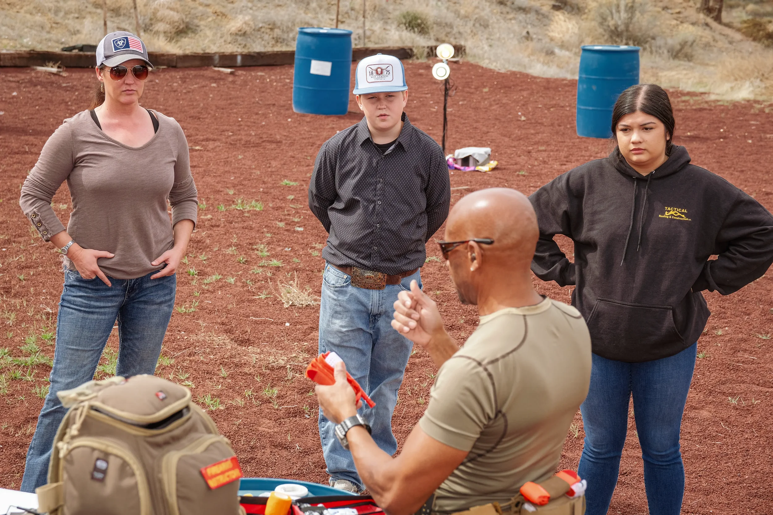 An outdoor setting with three women and one man. The man, who is bald and wearing sunglasses, is sitting and holding a small object. The women are standing around him, looking at him attentively. Two blue barrels are visible in the background on a re