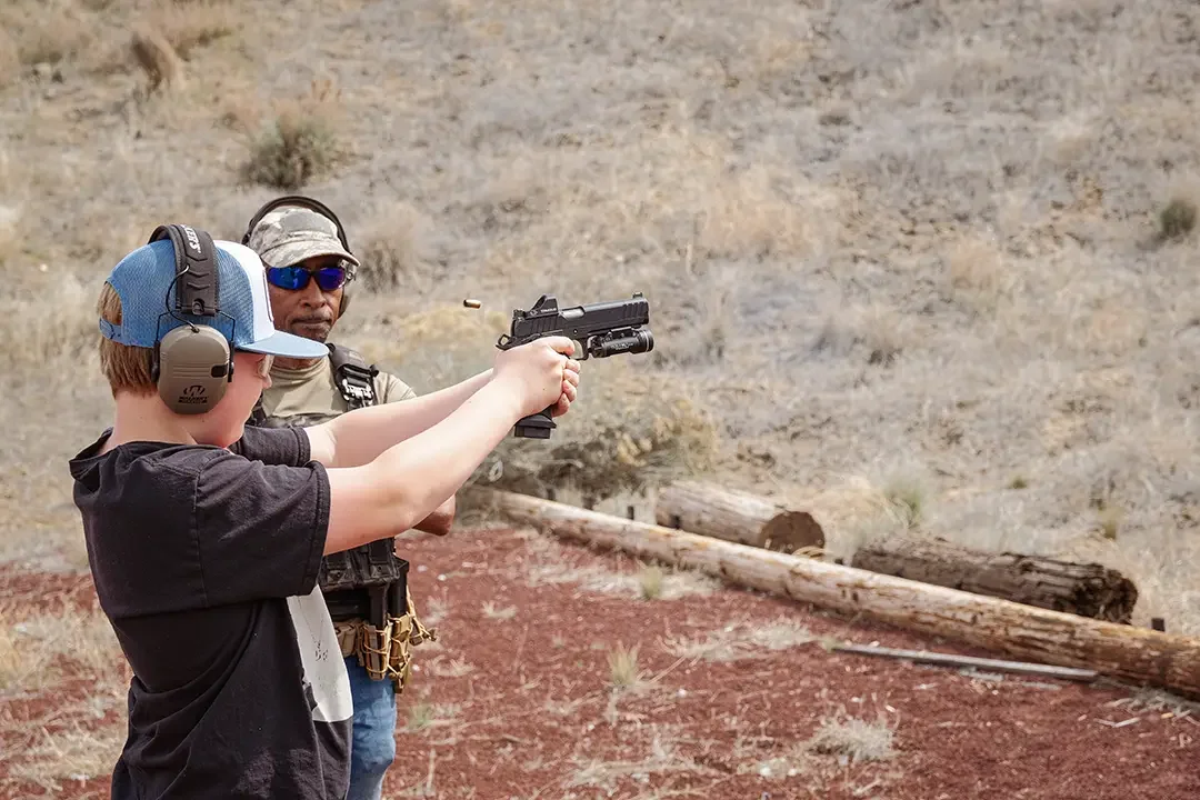 A young boy and an adult man at a shooting range, practicing or learning gun safety. The boy is holding a handgun, aiming it, while wearing hearing protection and a cap. The man, standing beside him, appears to be supervising or instructing, also equ