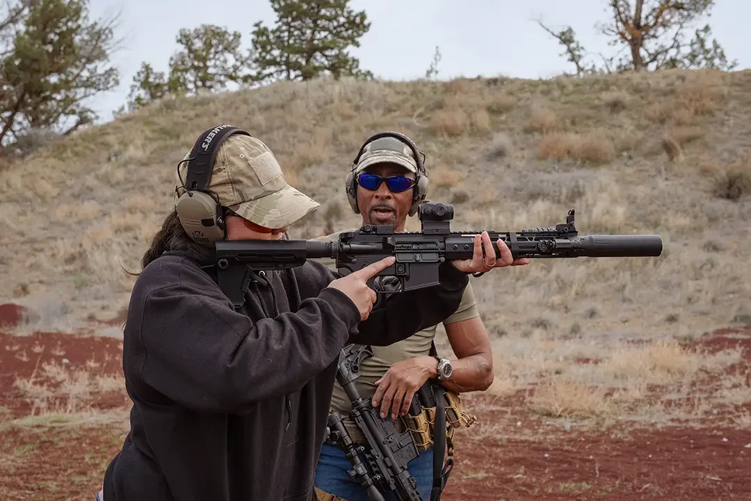 Two people at an outdoor shooting range with one holding a sniper rifle and the other instructing, both wearing tactical gear and headphones.