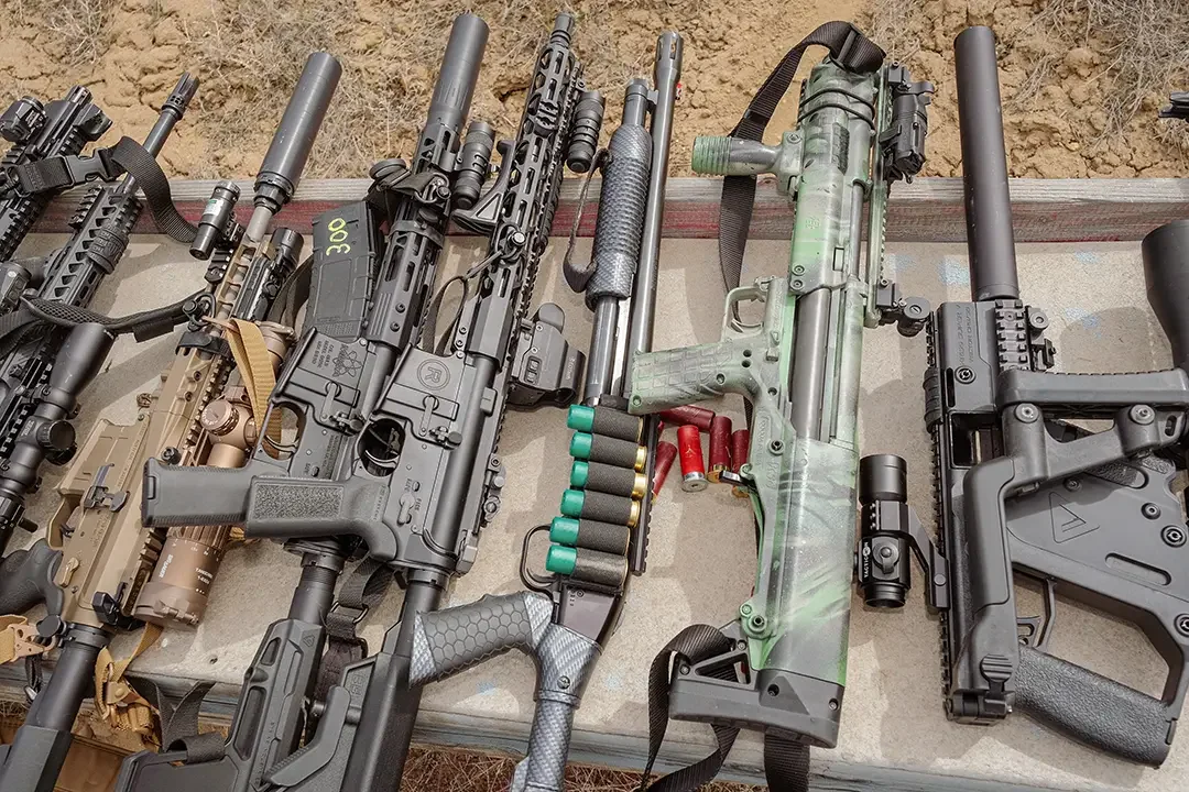 Various firearms including rifles and a shotgun laid out on a table with ammunition nearby.