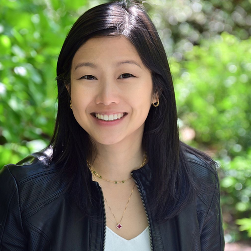 A young woman with dark hair smiling outdoors with green foliage in the background.