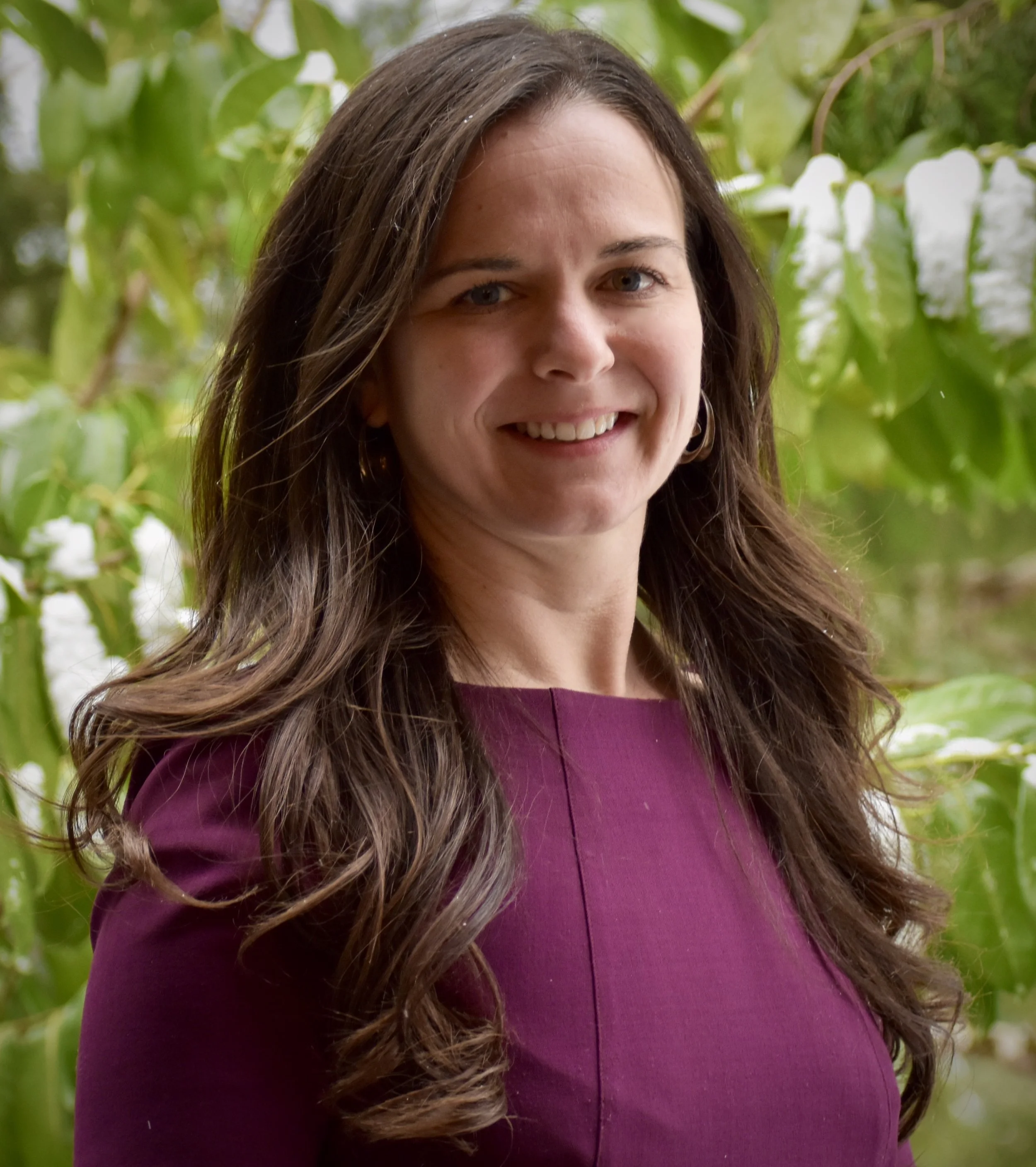 A woman with long brown hair and hoop earrings smiling outdoors, with green leafy plants in the background.