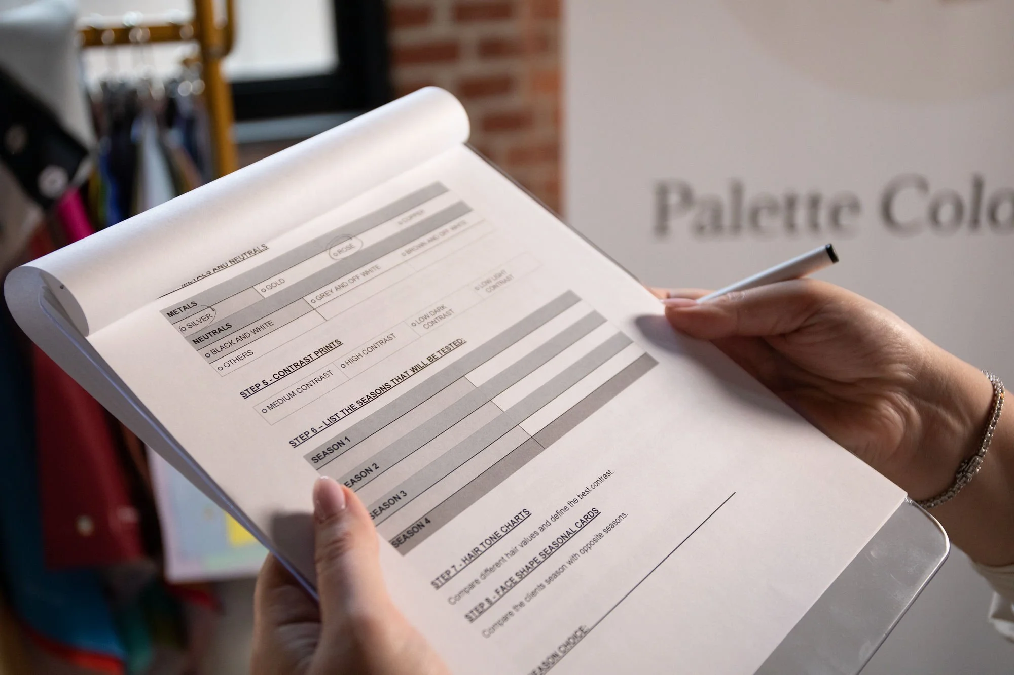 A person holds a checklist printed on white paper, with organized categories and steps related to color analysis and styling while standing indoors near a brick wall and a sign that says "Paletic Col" in the background.