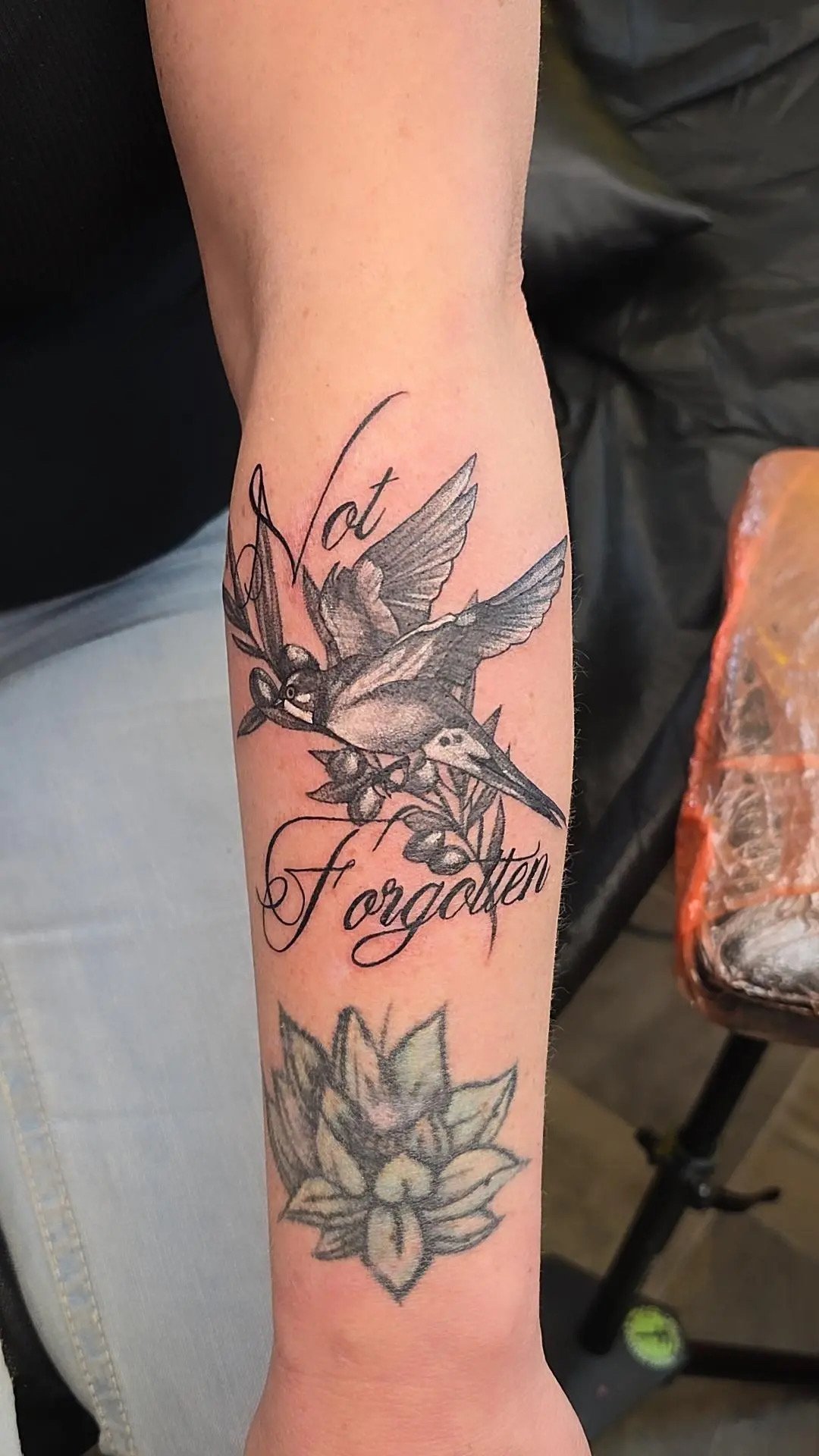 A tattoo of a hummingbird, with the words "Not Forgotten" written in cursive script, and a flower below.