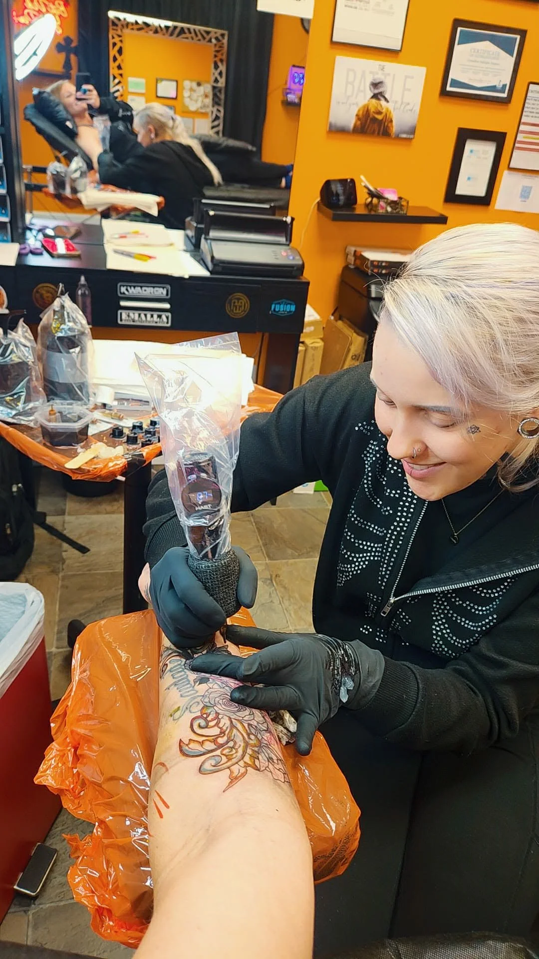 A woman with blonde hair getting a colorful tattoo on her arm in a tattoo shop, with various equipment and supplies visible on the table.