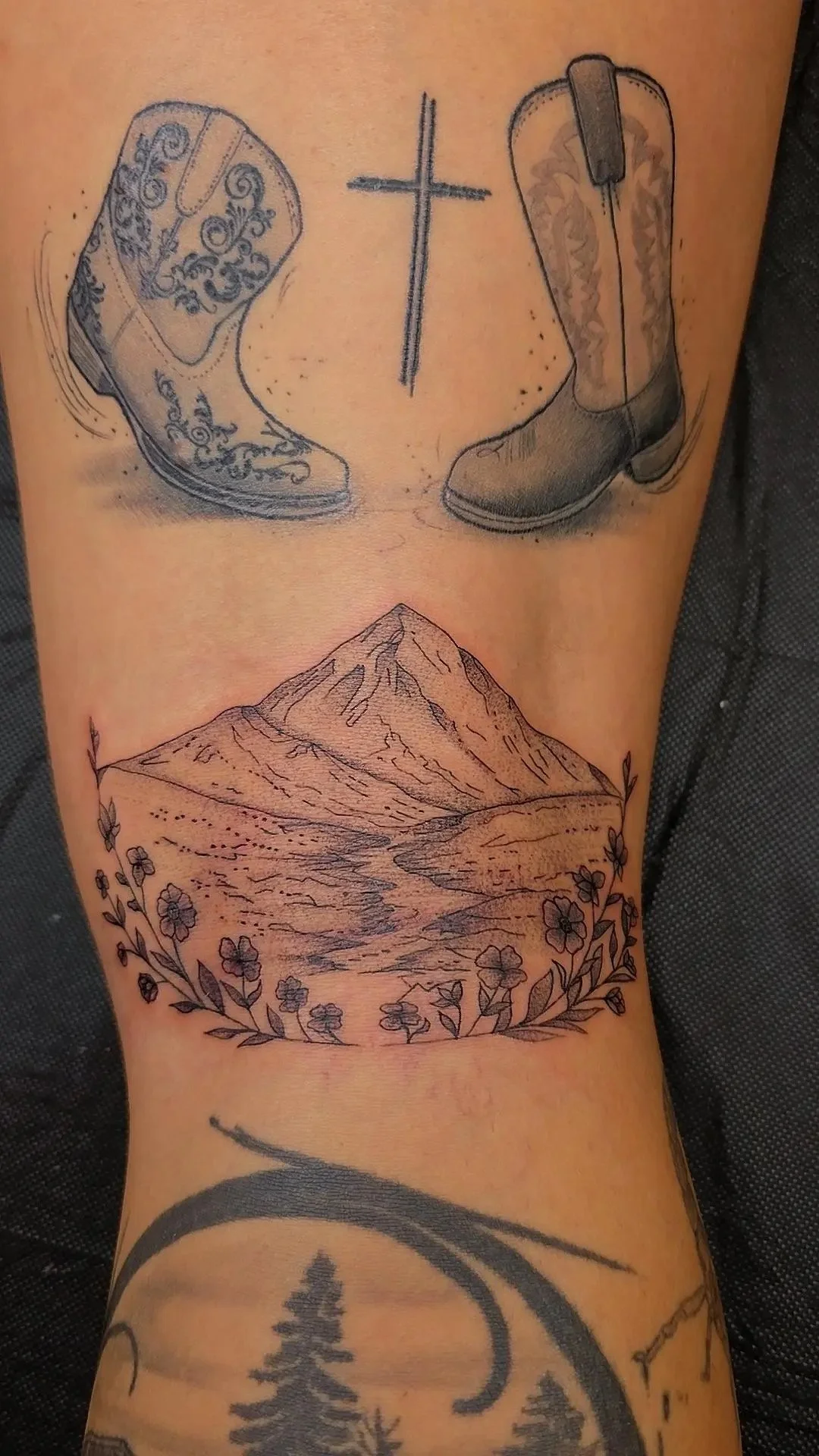 Tattoo of a mountain scene with flowers at the base, surrounded by other tattoos of cowboy boots and a cross, on a person's arm.