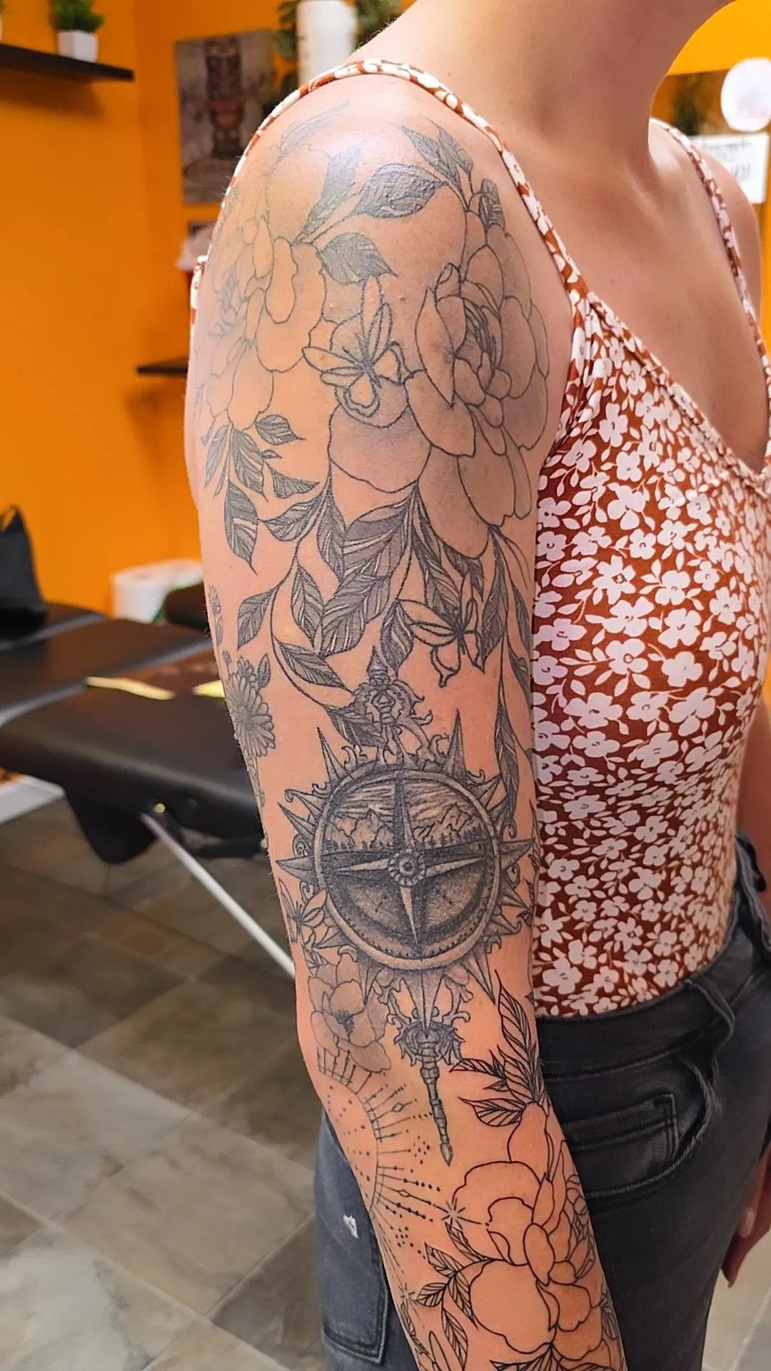 A woman with a floral tattoo sleeve on her upper arm, featuring large flowers, leaves, a compass, and a butterfly, wearing a floral-patterned tank top.