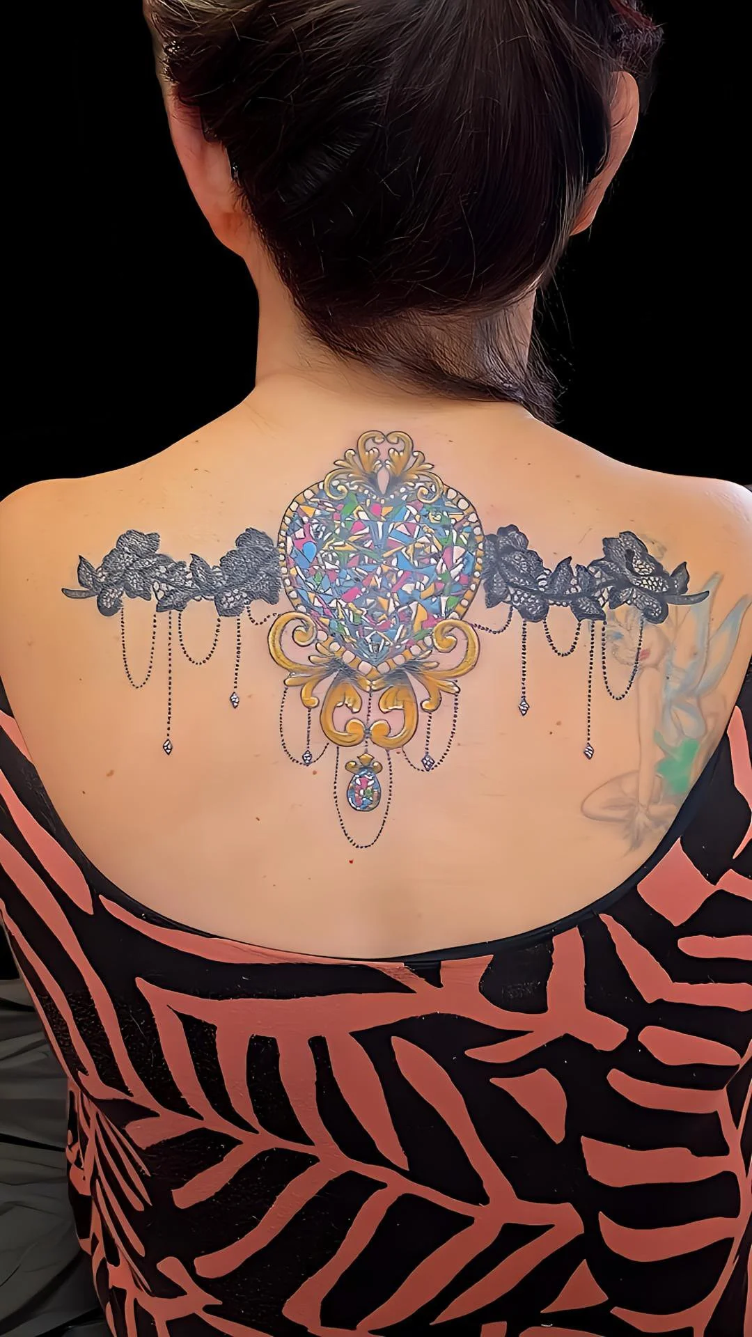 A woman with a colorful tattoo of a stained glass heart and ornate gold accents on her upper back, with black lace and dangling jewel designs, partially visible black and pink patterned dress.