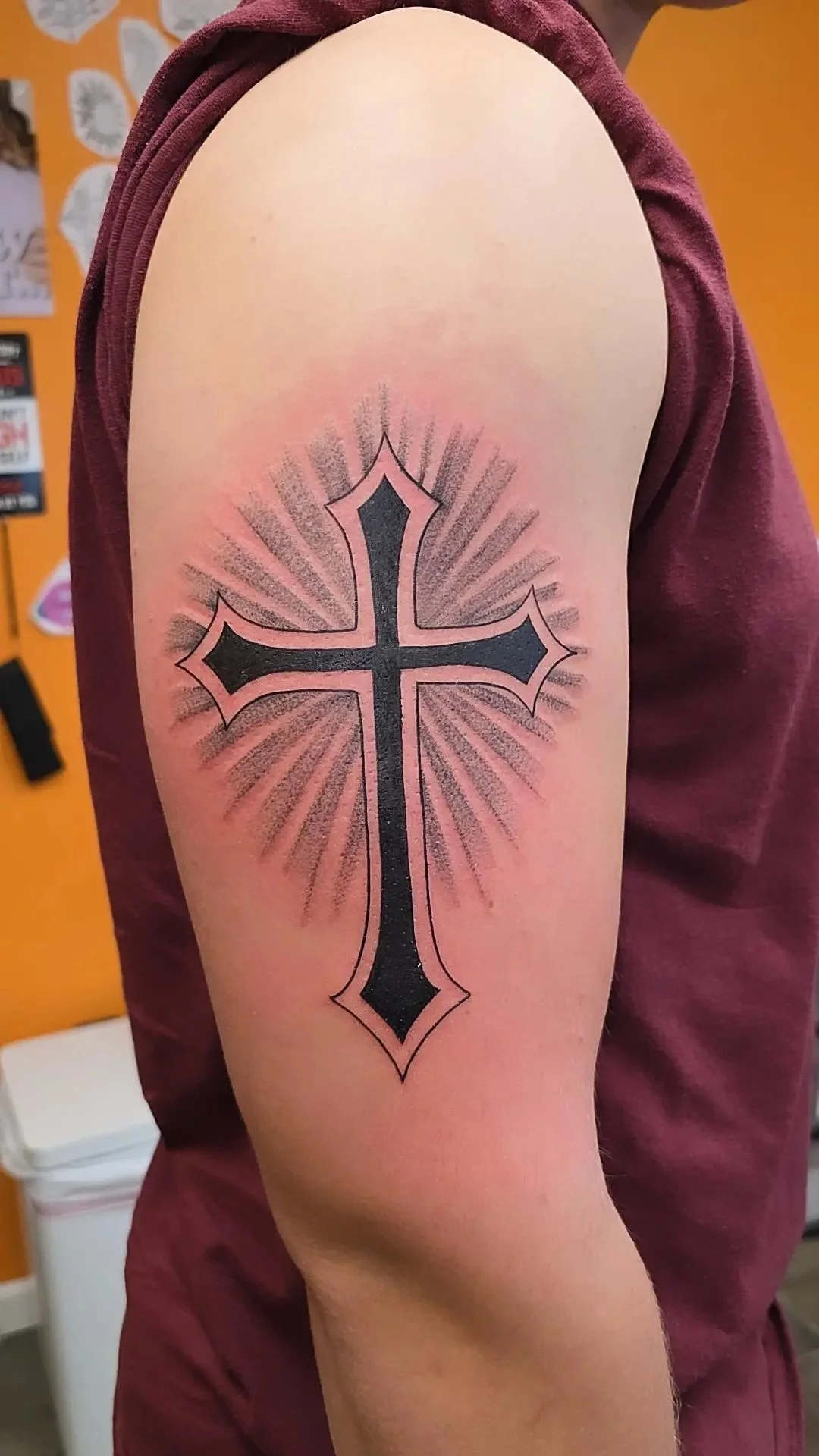 Tattoo of a black cross with pink outlines and radiating lines on the upper arm of a person wearing a maroon shirt.