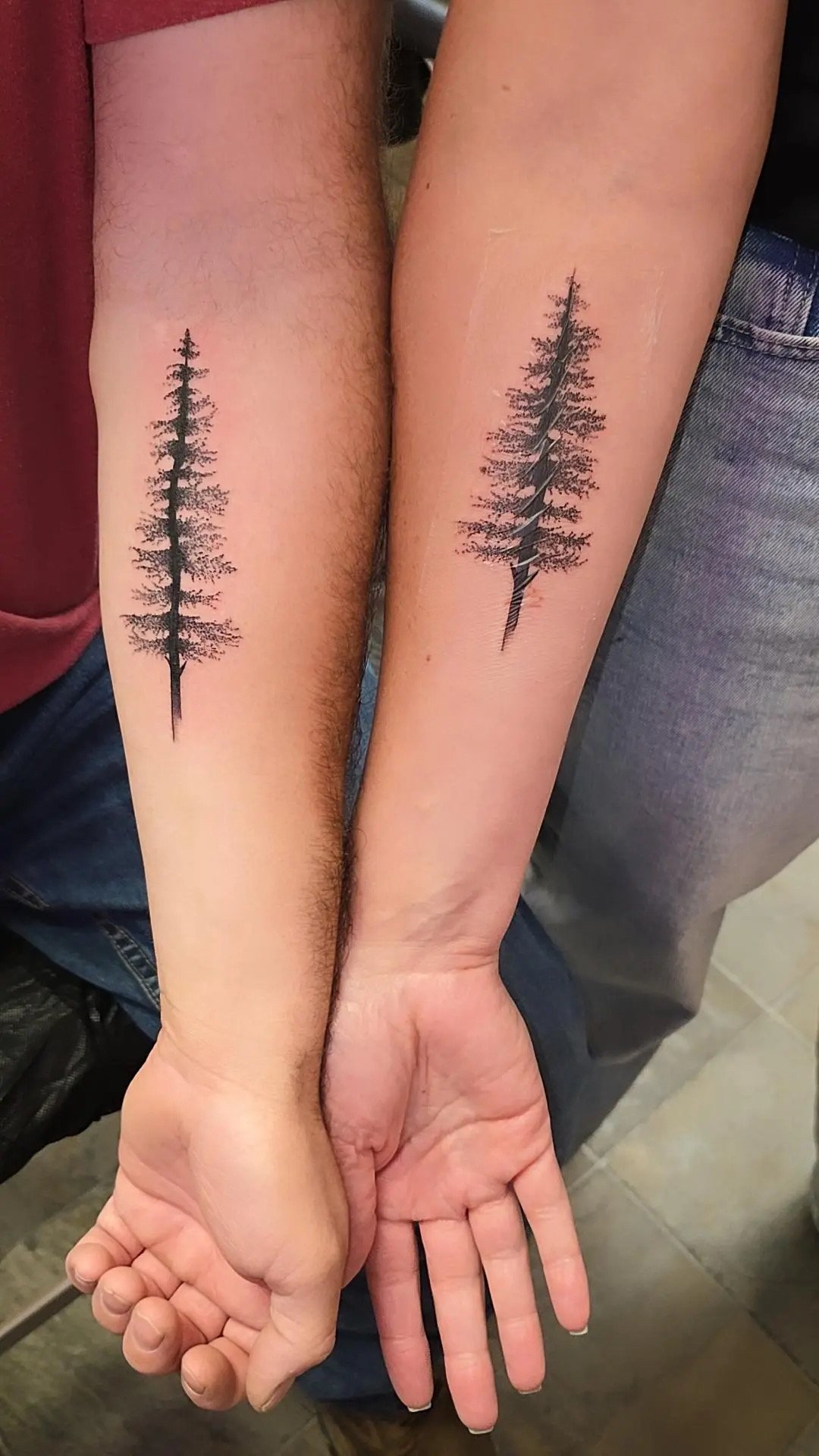 Two people standing side by side, each has a matching tattoo of a black silhouette of a pine tree on their inner forearm.