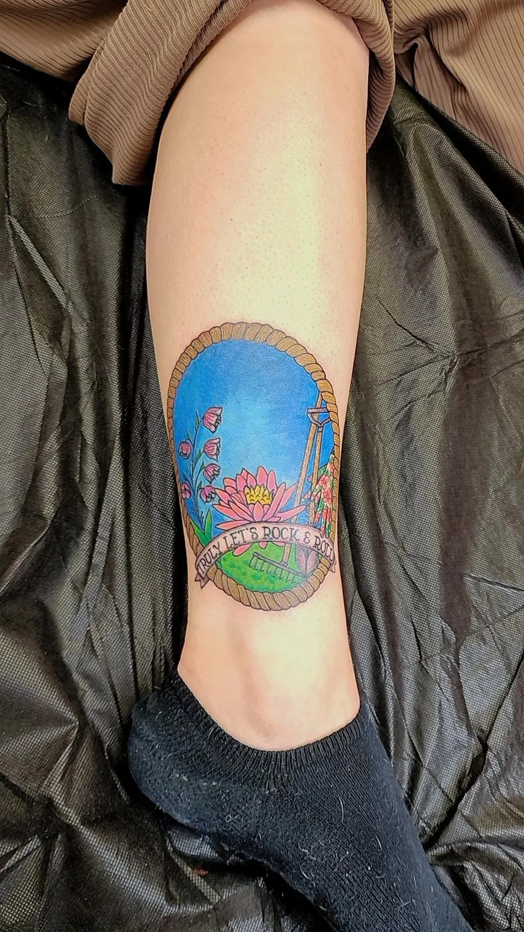 Colorful tattoo on person's leg of flowers, a swing, a blue sky, and a banner with the phrase "TRULY LET'S ROCK & ROLL."