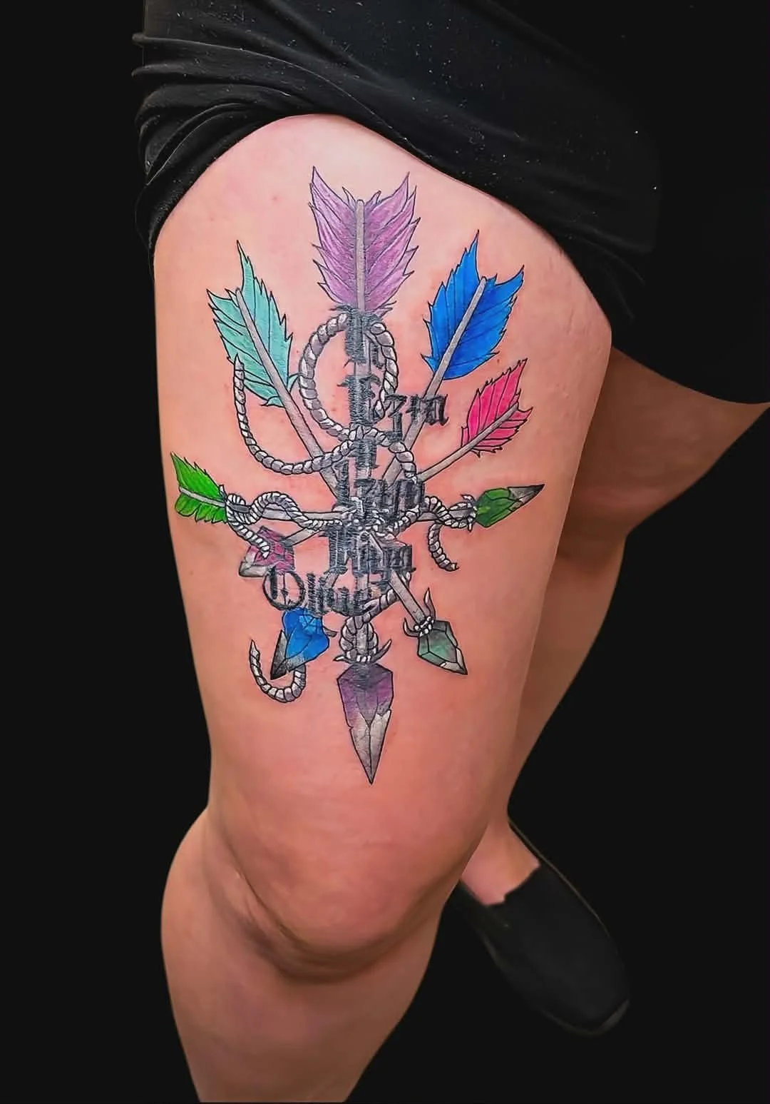 Colorful tattoo on thigh featuring a knife and arrow intertwined with multicolored feathers.
