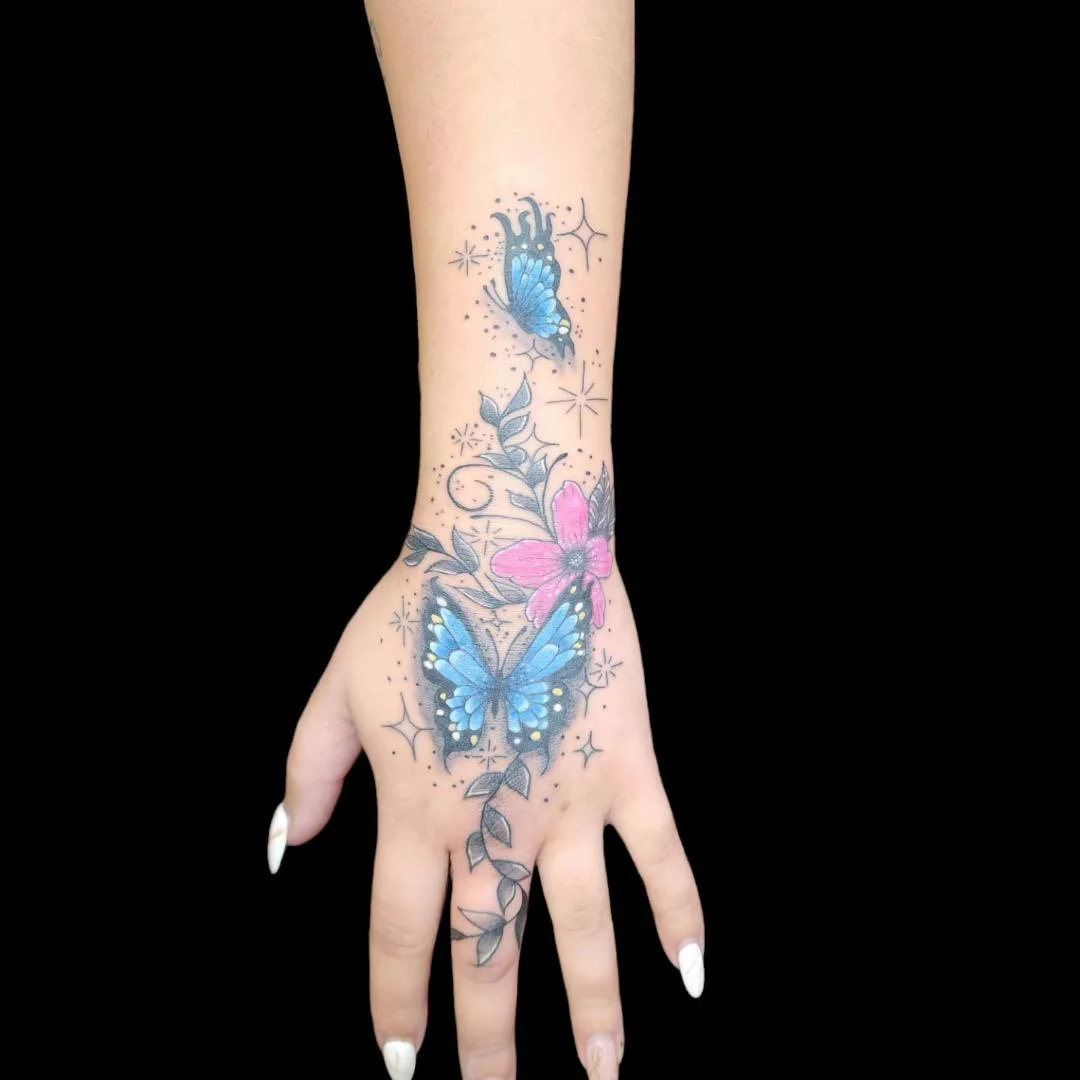 Tattoo of two blue butterflies, pink flower, and leafy vines on a person's hand and wrist.