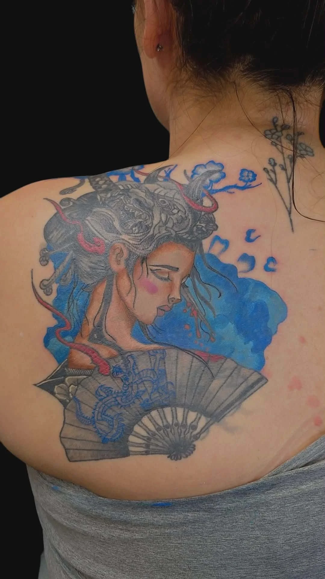 Colorful tattoo of a woman's profile with flowing hair, holding a fan, with a blue background and flower details on the shoulder.