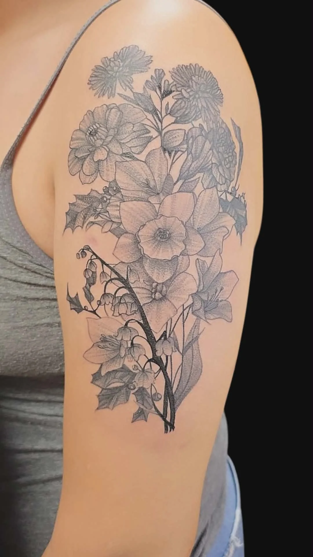 Black and gray floral tattoo on person's upper arm featuring various flowers, leaves, and holly accents.