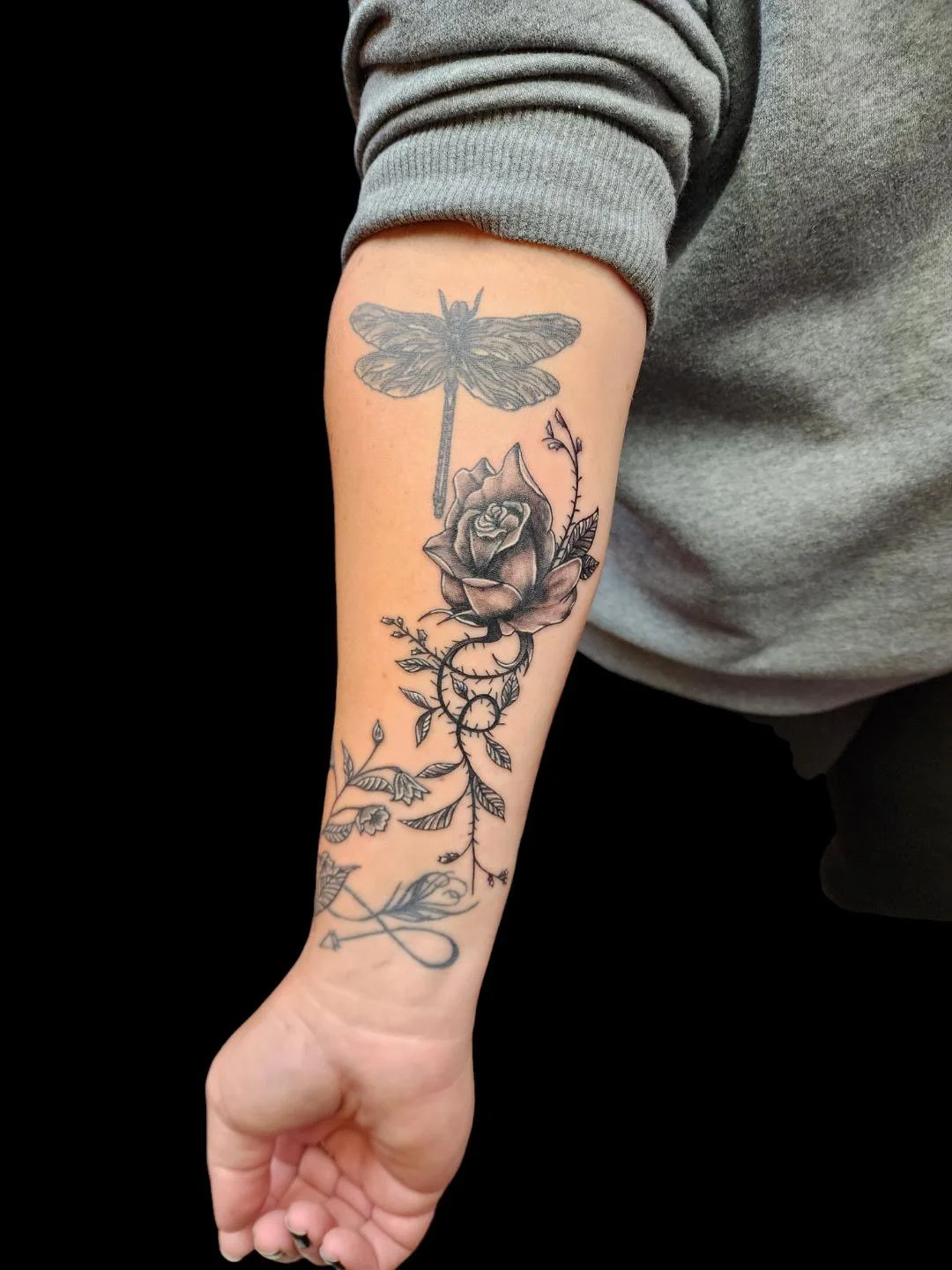 Black and gray tattoo of a dragonfly above a rose with leaves and thorny vines on a person's forearm.