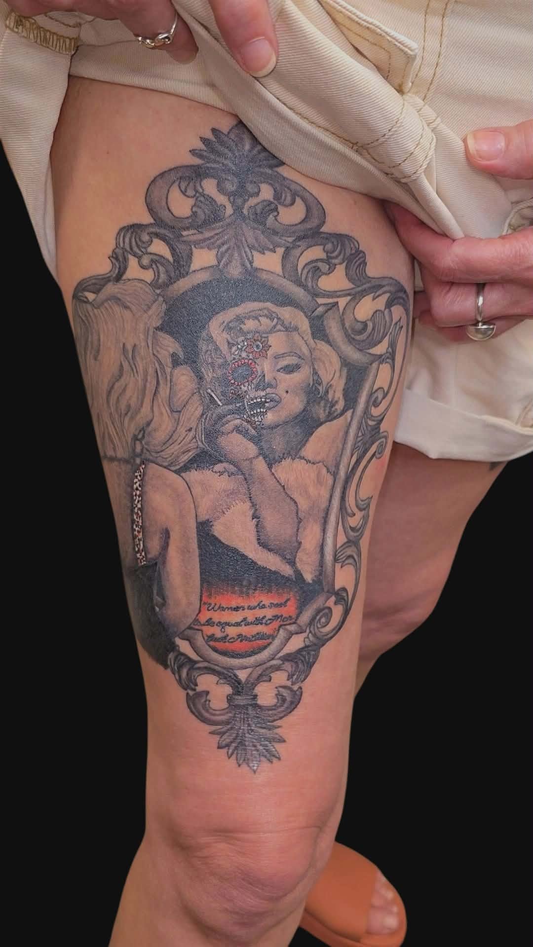 Tattoo of a vintage-style portrait of a woman with a skull face, framed by ornate scrollwork and floral designs, with a red and black banner containing text at the bottom.