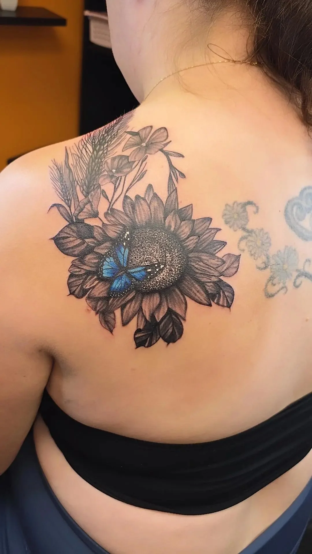 Close-up of a person's shoulder and upper chest with a large, detailed sunflower tattoo. The tattoo features a blue butterfly perched on the sunflower.