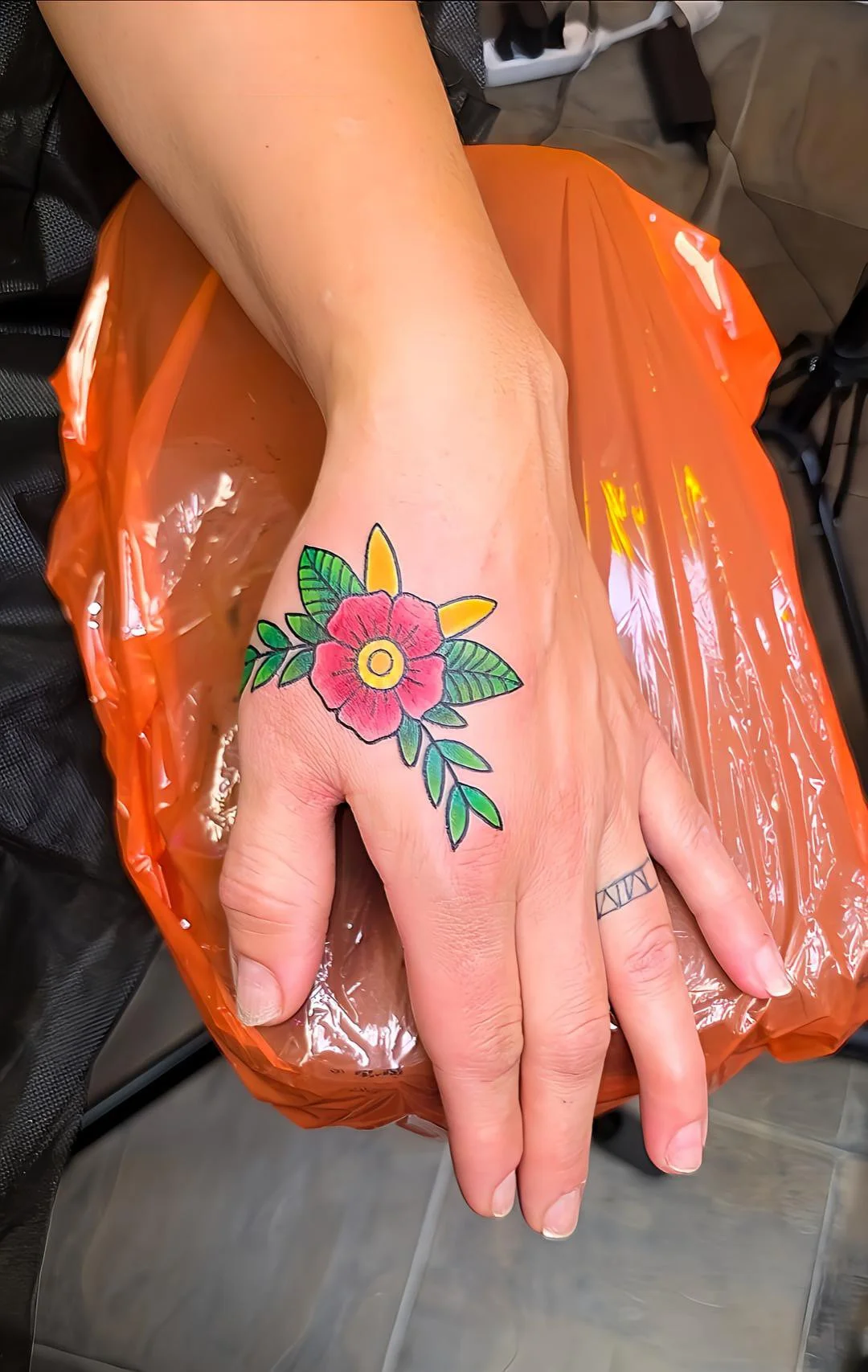A person's hand with a colorful tattoo of a red flower and green leaves resting on an orange plastic surface.