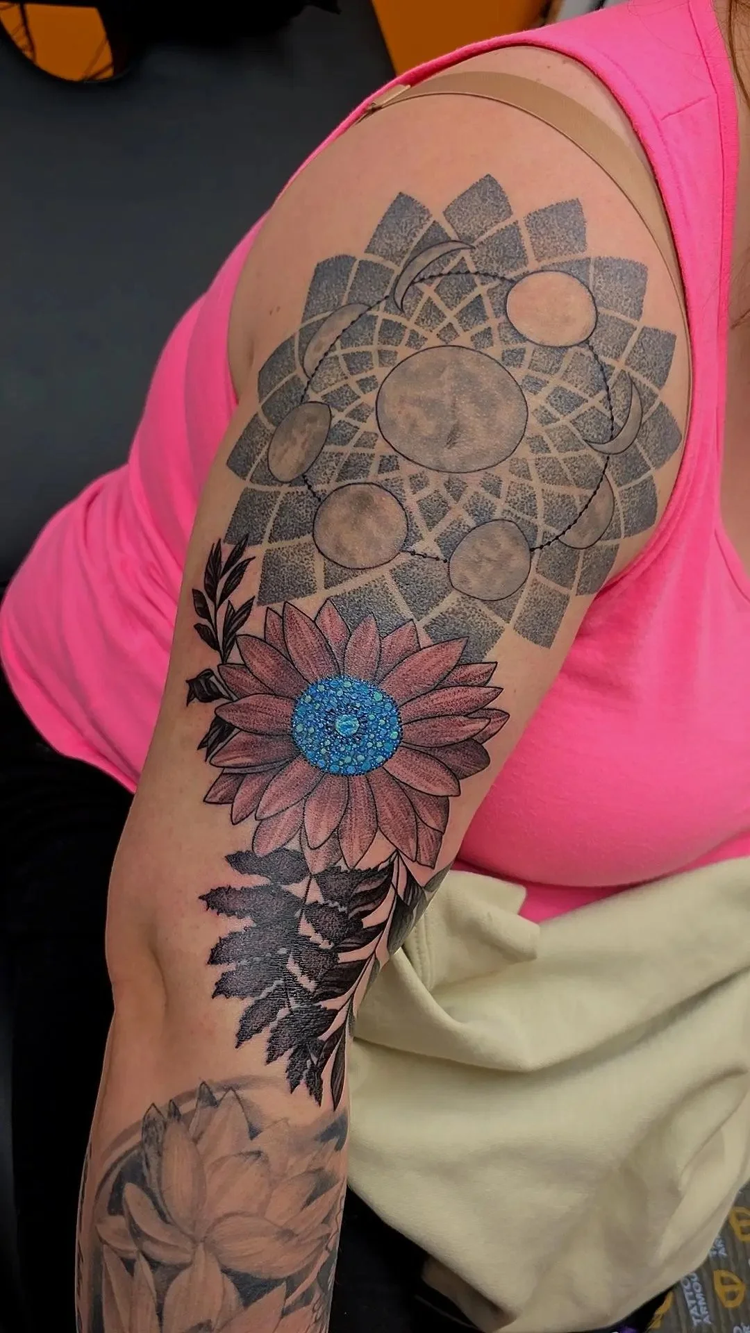 A woman with a shoulder tattoo featuring a geometric moon phase pattern, a large sunflower with blue center, and additional floral and leaf designs.