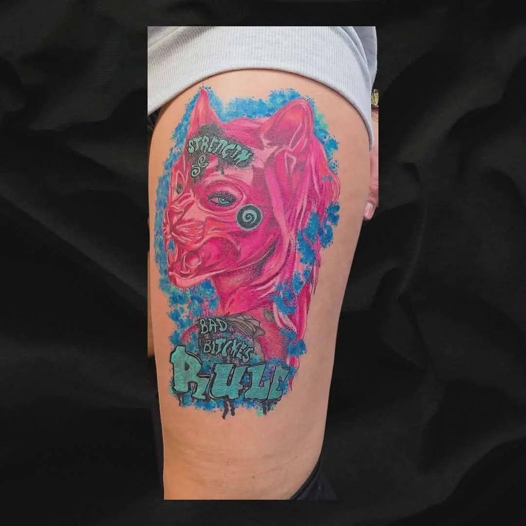 Colorful tattoo of a pink wolf with blue and gray details, surrounded by graffiti-style text that reads 'Strong,' 'Bad Bitch,' and 'Queen' on a person's thigh.