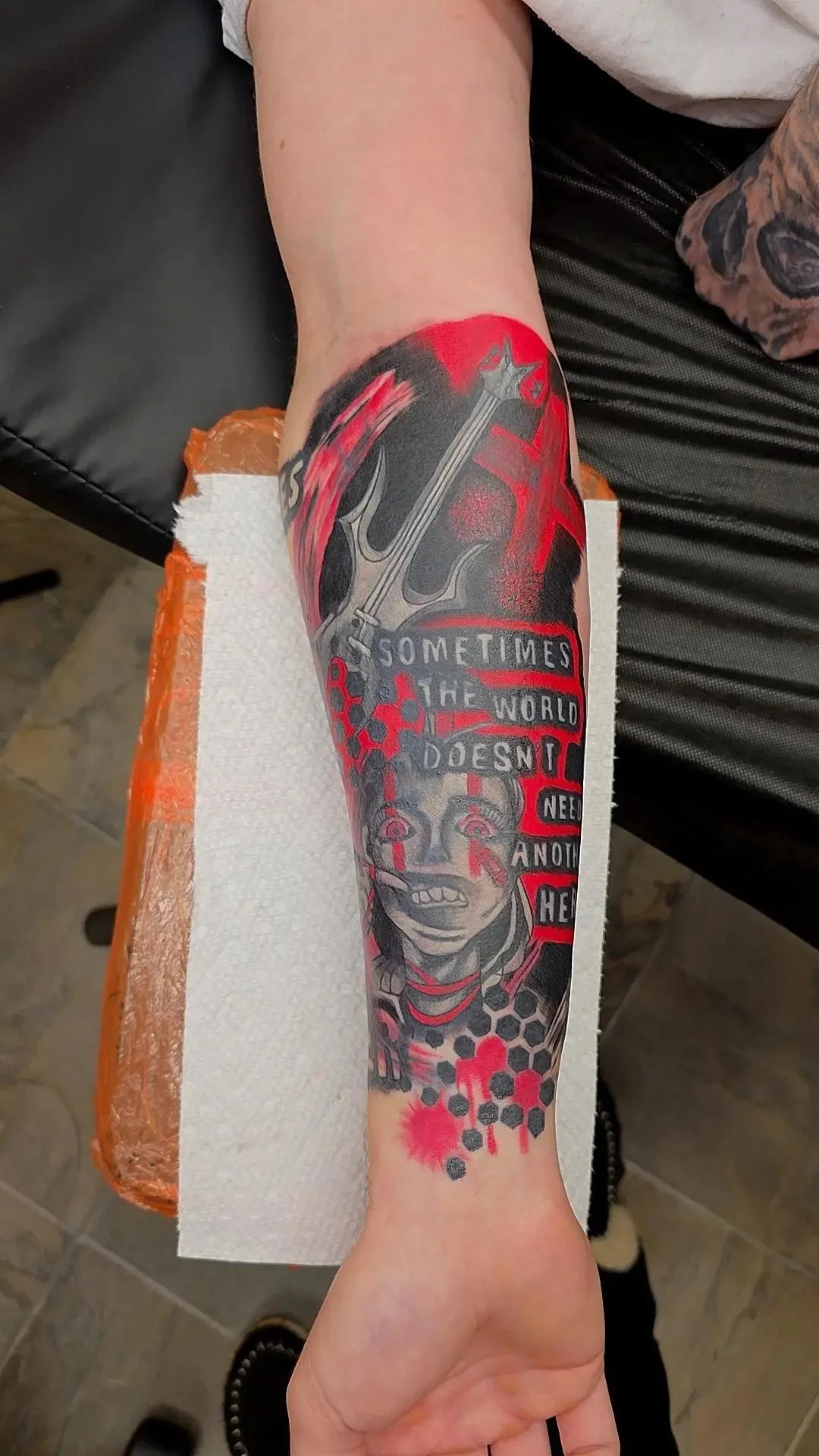 Tattoo on person's forearm featuring a woman with beaded neck adornments, a background of black and red, a large trident, and text that reads 'Sometimes the world doesn't need another hero'.