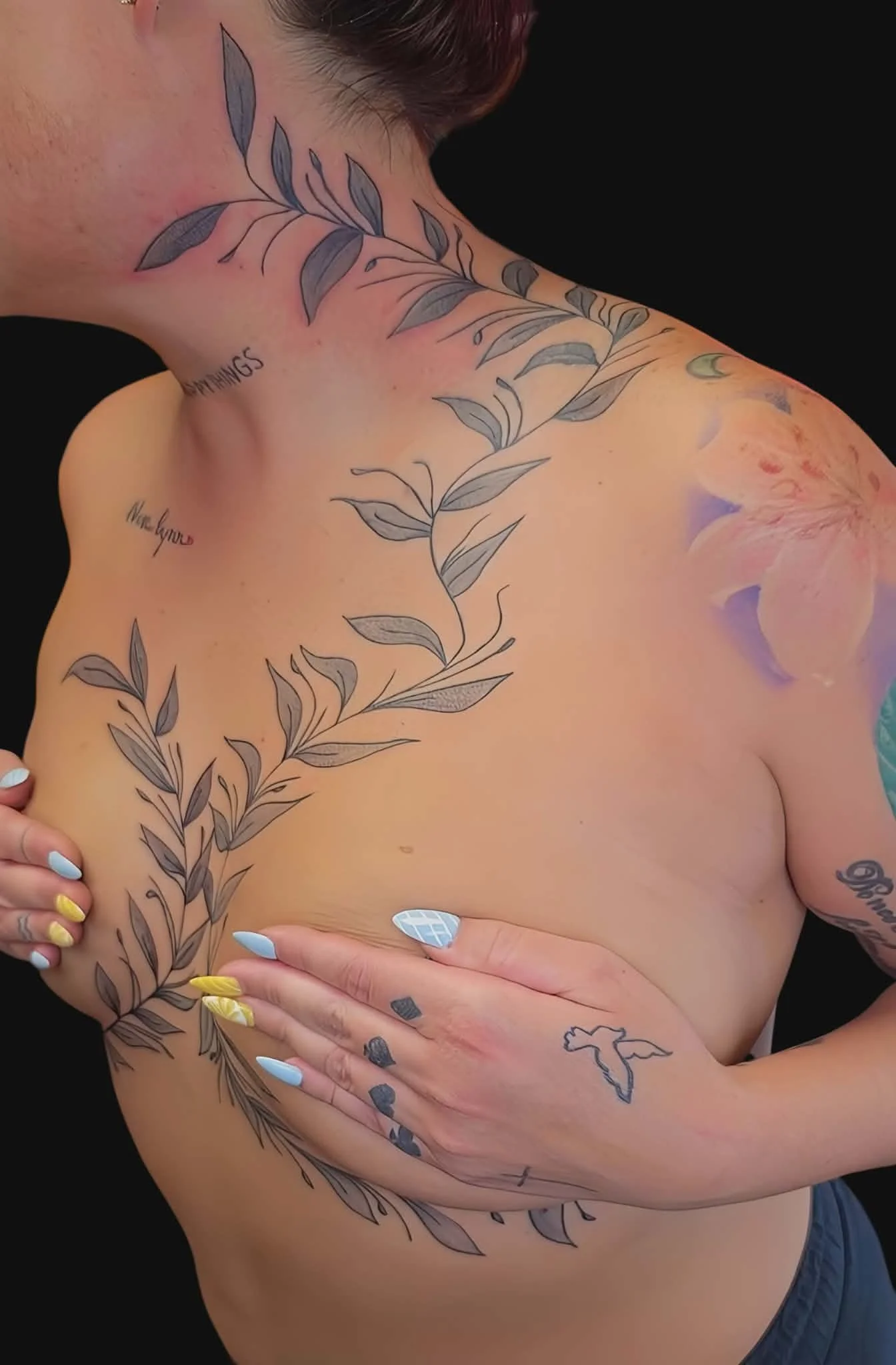 Person with multiple tattoos, including leaf designs on shoulder and chest, bird outline on hand, and various smaller tattoos, holding their breast with their hand, against a black background.