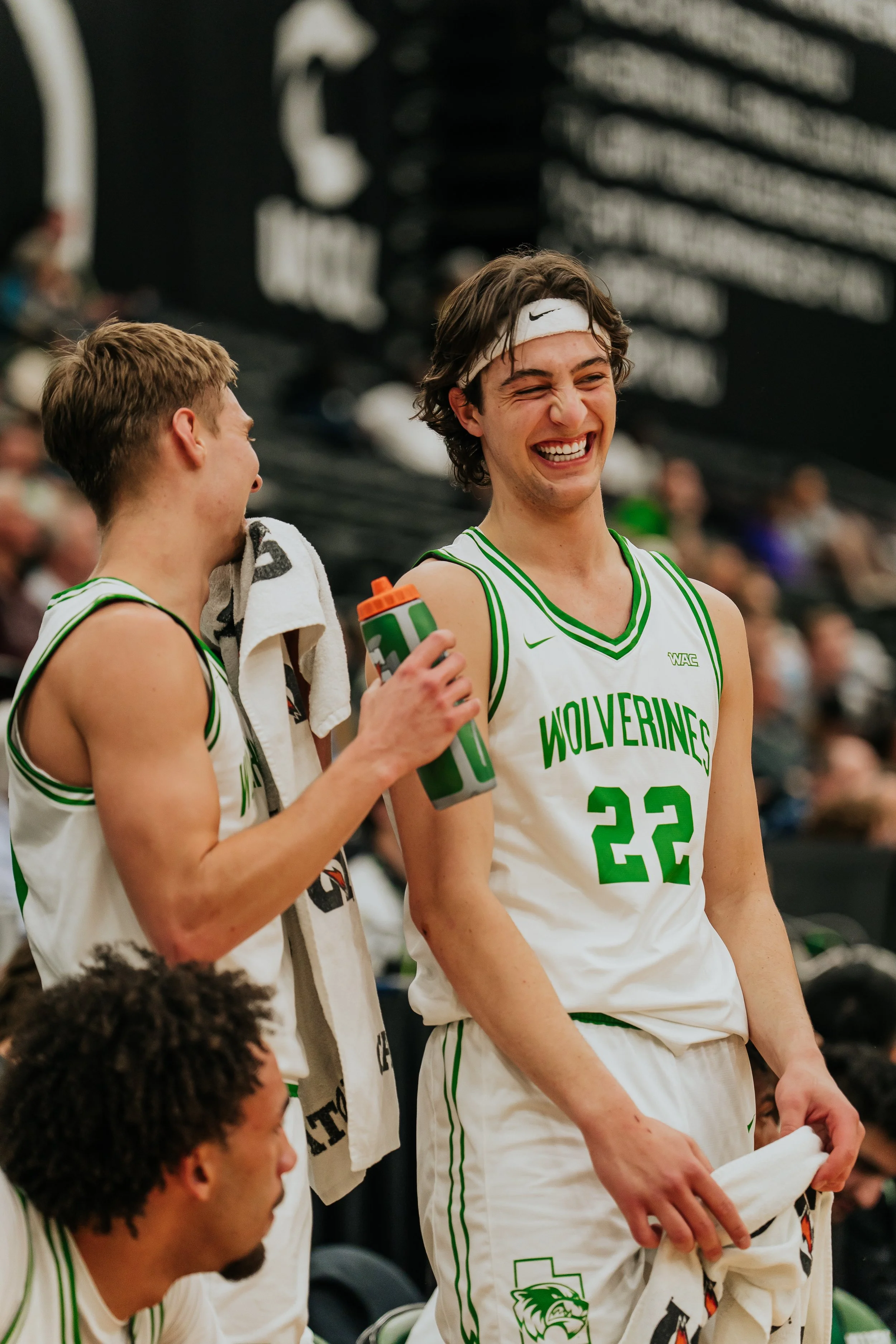 UVU Men's Basketball player laughs with their teammate while sitting on the side lines. 