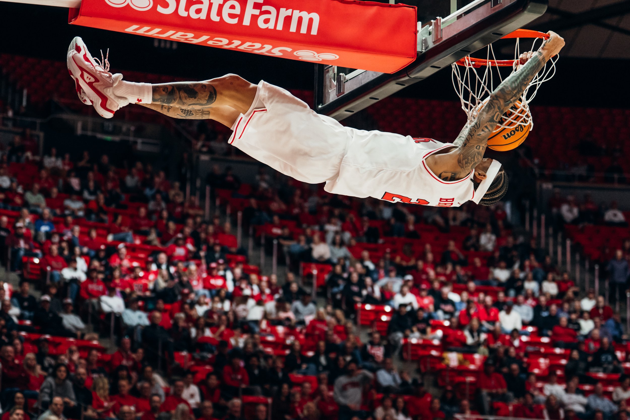 A basketball player with tattoos and wearing a white jersey dunks a basketball into the hoop at an indoor arena, with a crowd watching in the background.