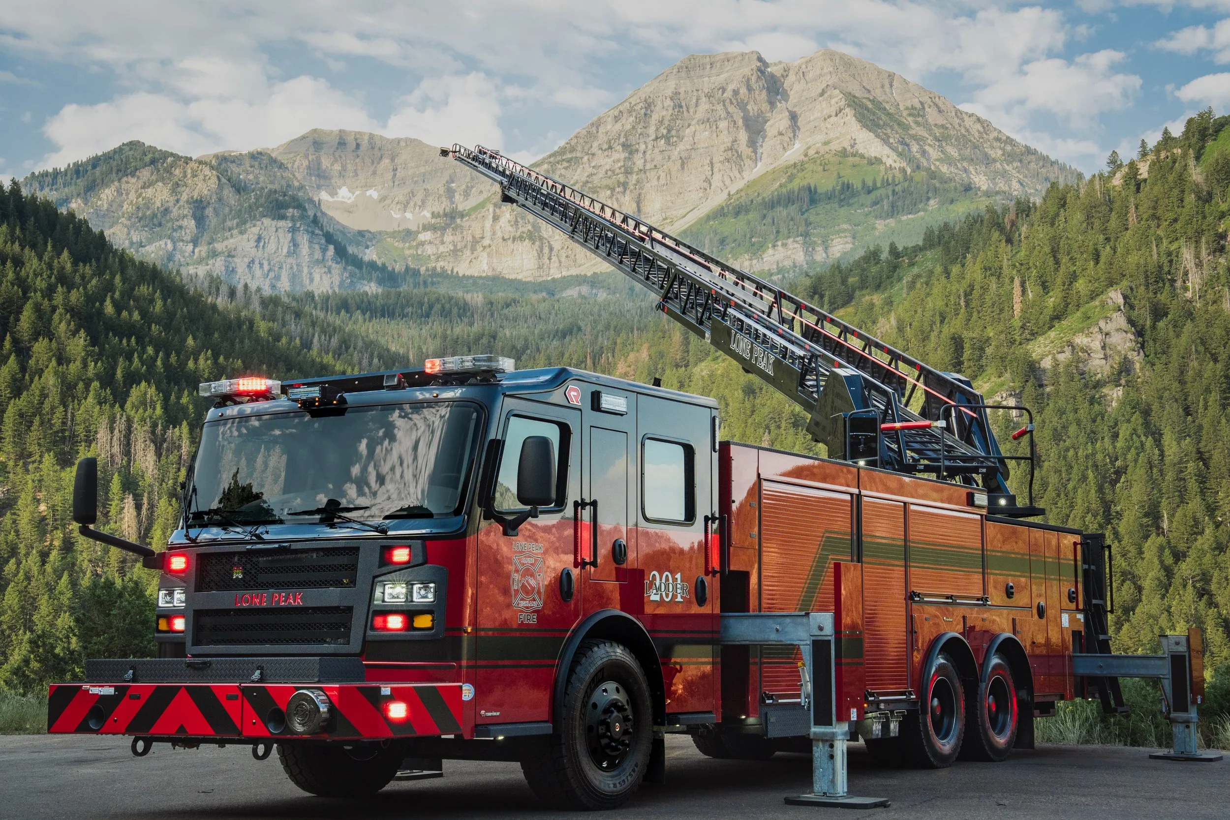 A red fire truck with an extended ladder and emergency lights on top, parked outdoors against a mountainous landscape with trees and a partly cloudy sky.