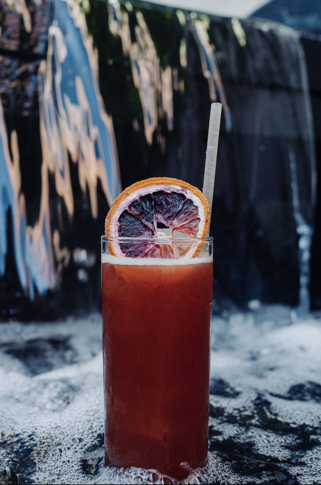 A tall glass of red cocktail with a slice of blood orange on the rim and a straw, set against a background of a waterfall with graffiti.
