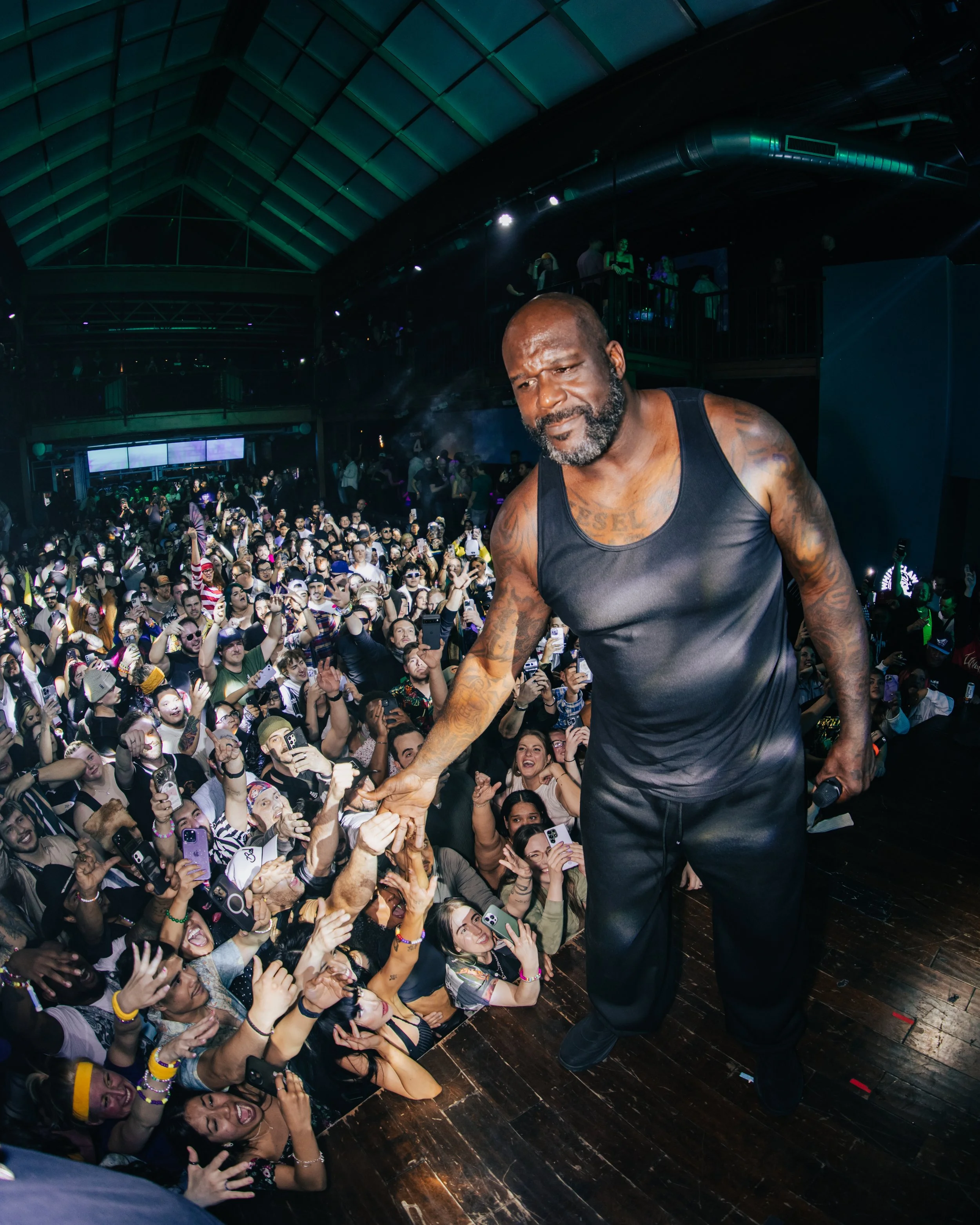 A man with a beard and tattoos on his arms, wearing a black tank top and black pants, is on a stage engaging with a large crowd at a concert or event inside a large, industrial-style venue with a high, arched ceiling. The audience is smiling, taking photos, and reaching out towards him.