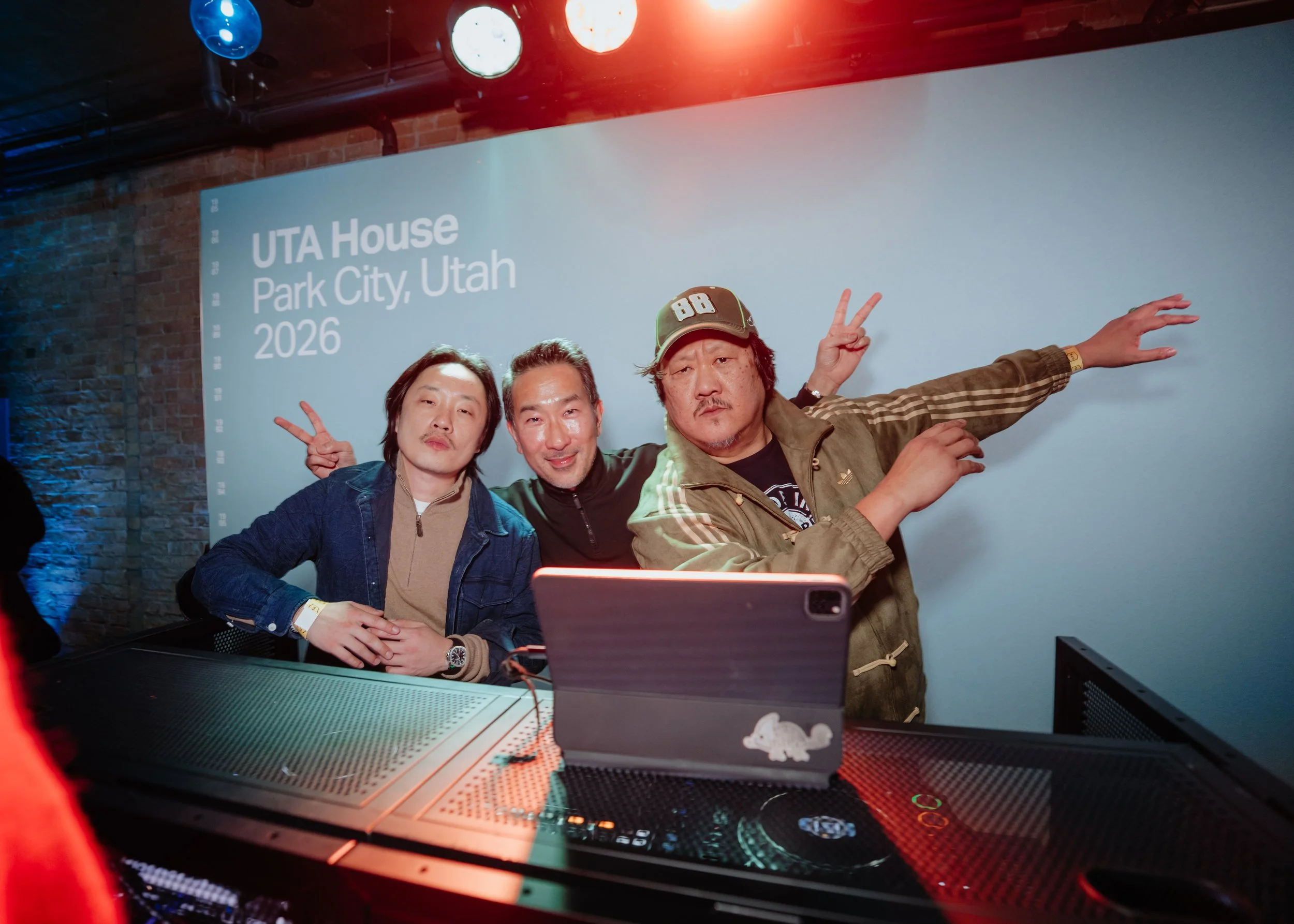 Three men taking a selfie in front of a presentation screen that reads 'UTA House, Park City, Utah, 2026.' They are posing with peace signs and smiling behind the DJ booth, with lights in the background.