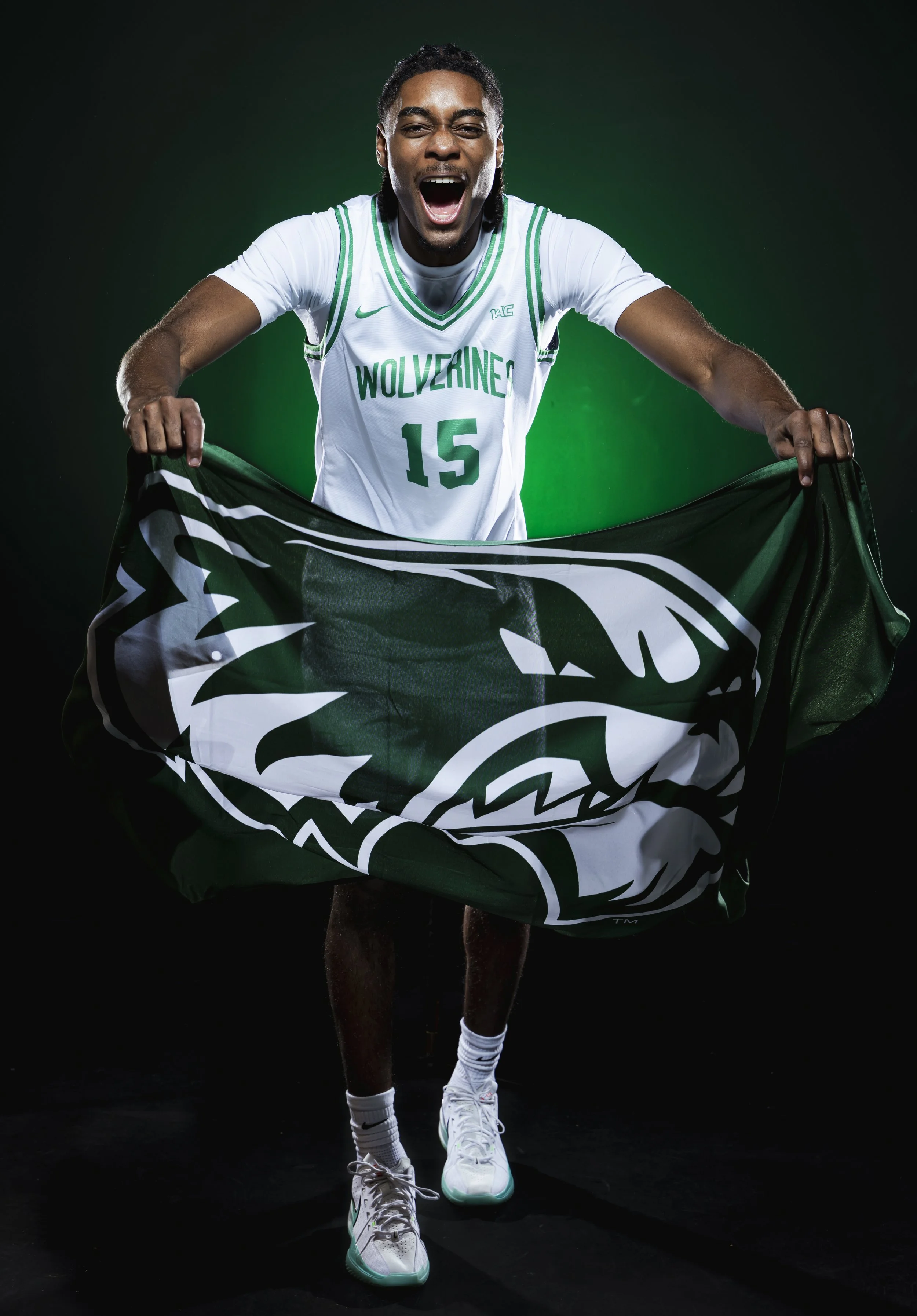 Young African American male basketball player in white jersey with green accents, number 15, holding a green and white Wolverines flag, against black background.